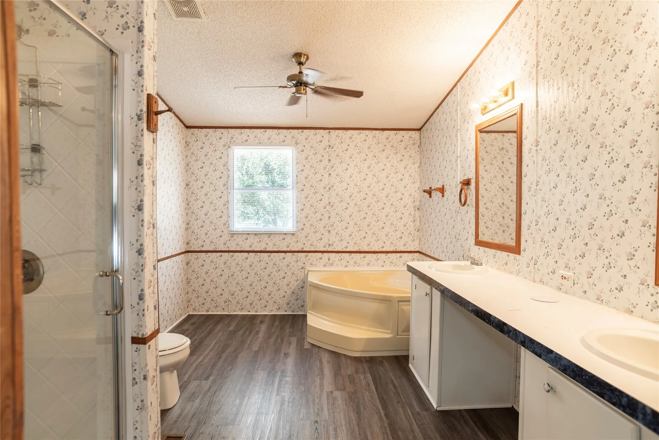 Bathroom featuring a corner soaking tub, dual vanity with undermount sinks, walk-in shower with glass enclosure, wood-finish flooring, and a ceiling fan