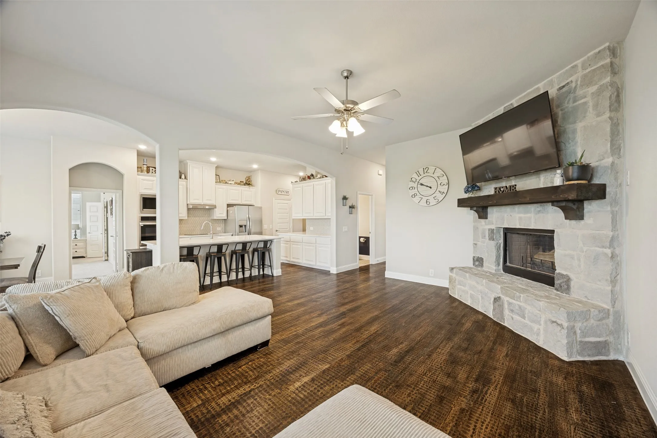 Spacious living area featuring a stone-clad fireplace with a substantial wood mantel, wood-finish flooring, multiple large windows with blinds, and a ceiling fan