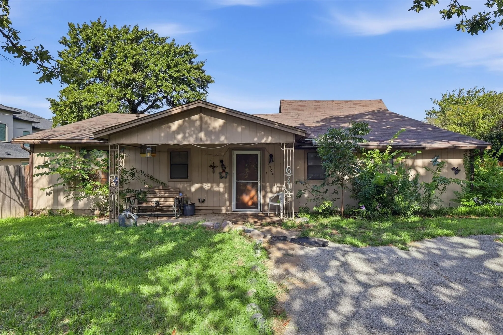 Front exterior featuring tan siding, a brown shingle roof, a covered porch entry, and mature trees surrounding the property