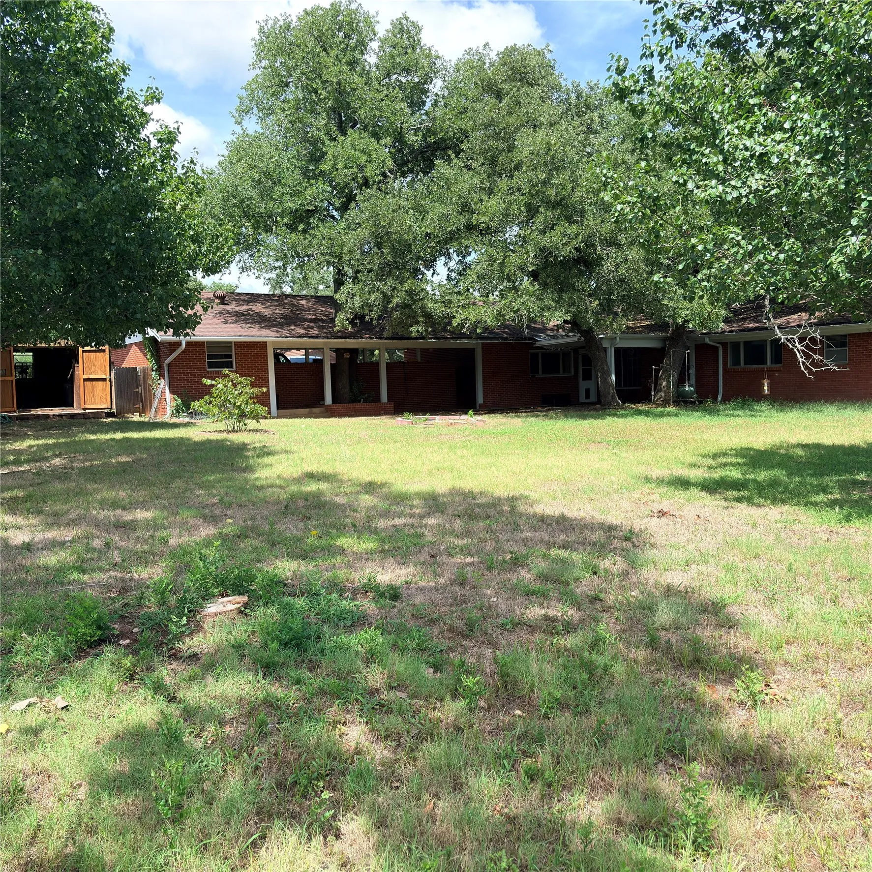 Expansive backyard with a brick-clad residence featuring a covered patio, a separate outbuilding with wooden doors, and numerous mature trees providing shade
