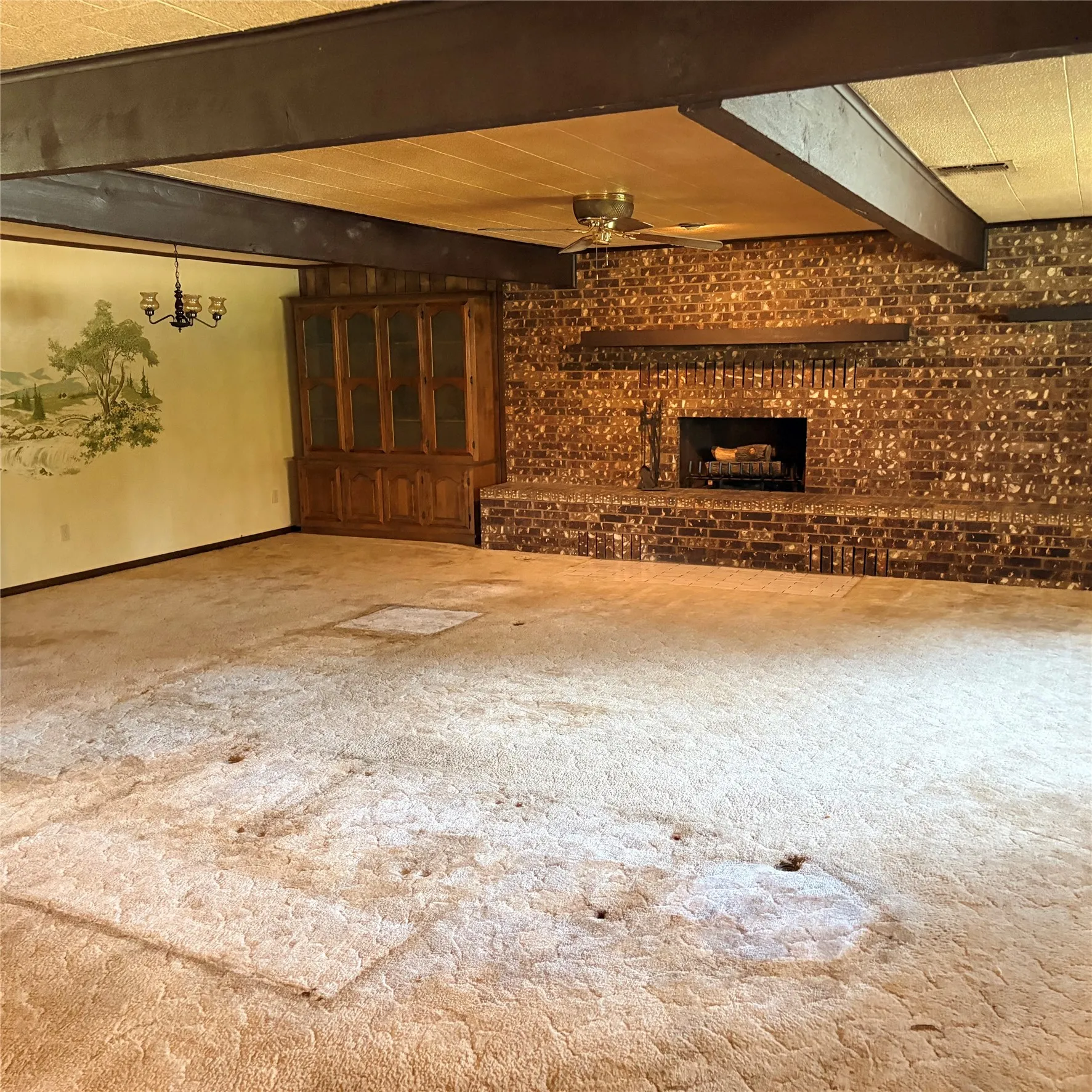 Spacious interior featuring exposed dark wood beams, a prominent brick fireplace with hearth, a built-in wood cabinet with glass doors, and a ceiling fan