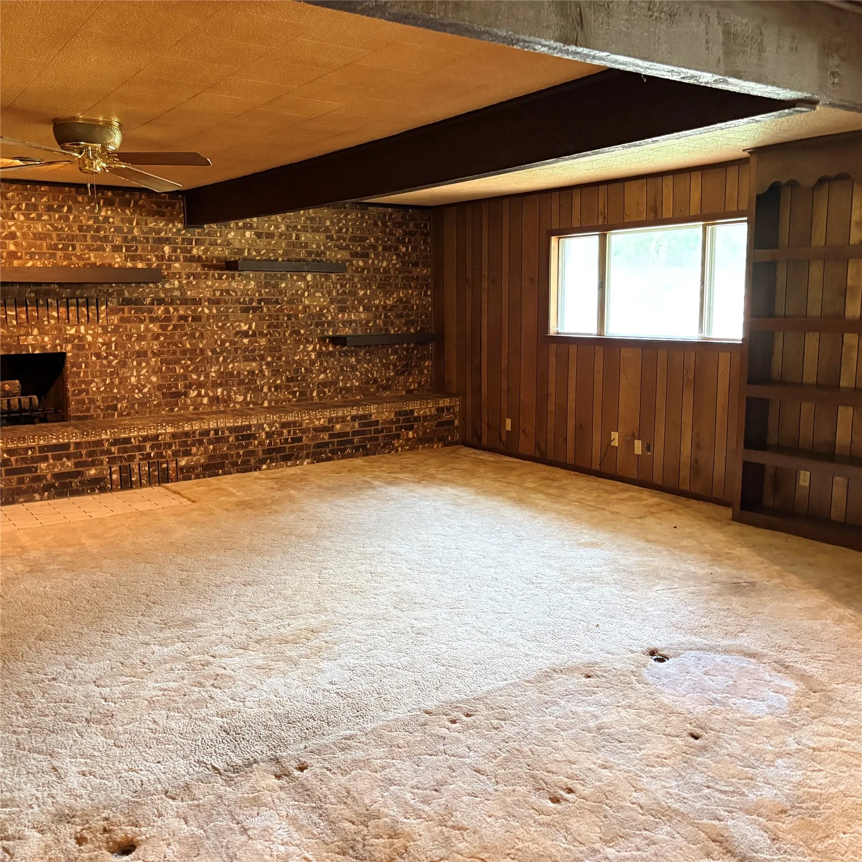 Spacious room featuring a brick fireplace with a raised hearth, wood-paneled walls, built-in shelving, and a ceiling fan