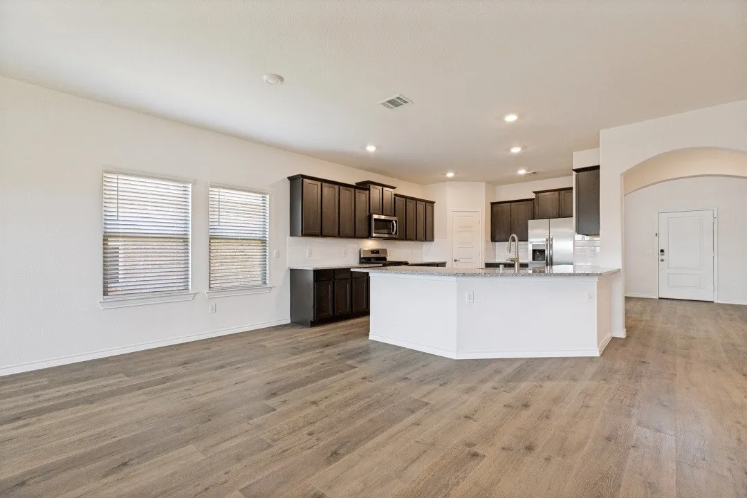 Open-concept living space with wood-finish flooring, a kitchen island, dark wood cabinetry, stainless steel appliances, and two windows with blinds
