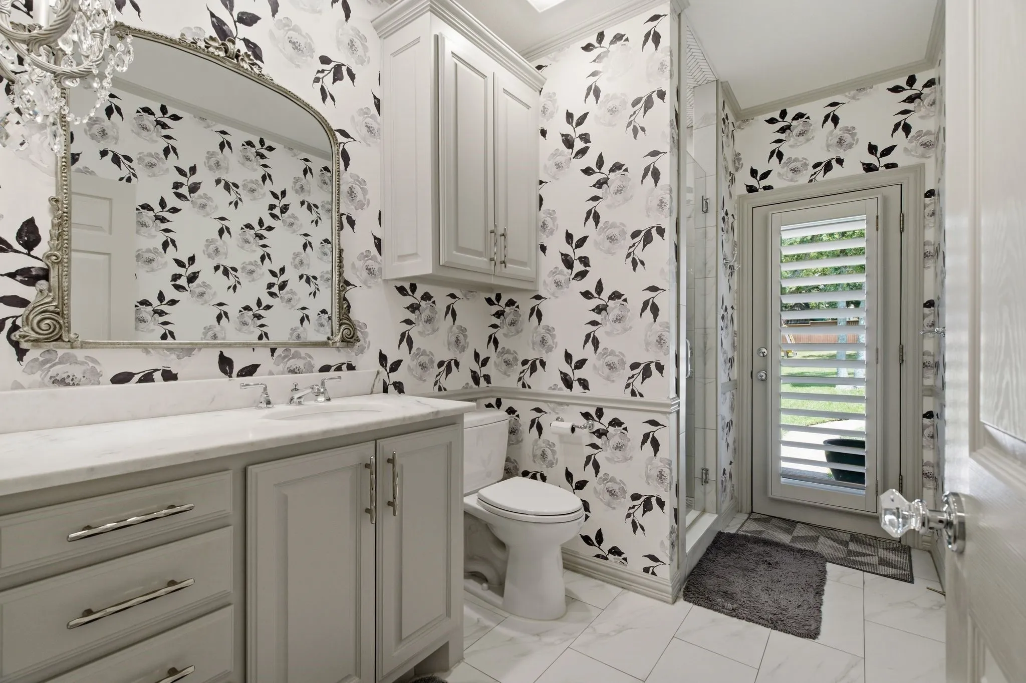 Bathroom featuring floral wallpaper, white vanity with stone countertop, ornate framed mirror, glass-enclosed shower, and exterior door with shutter blinds