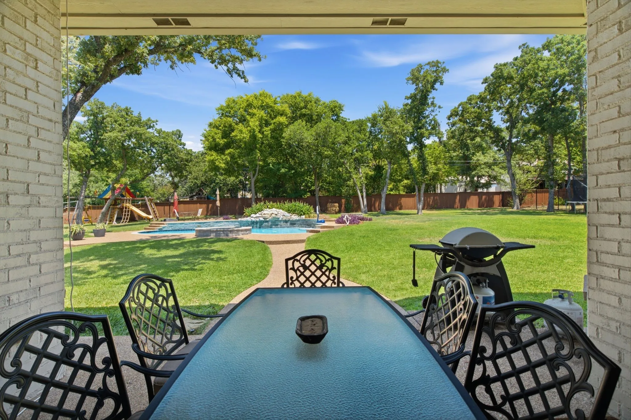 Covered patio overlooking a spacious backyard featuring a freeform swimming pool, integrated spa with rock waterfall, expansive lawn, and mature trees