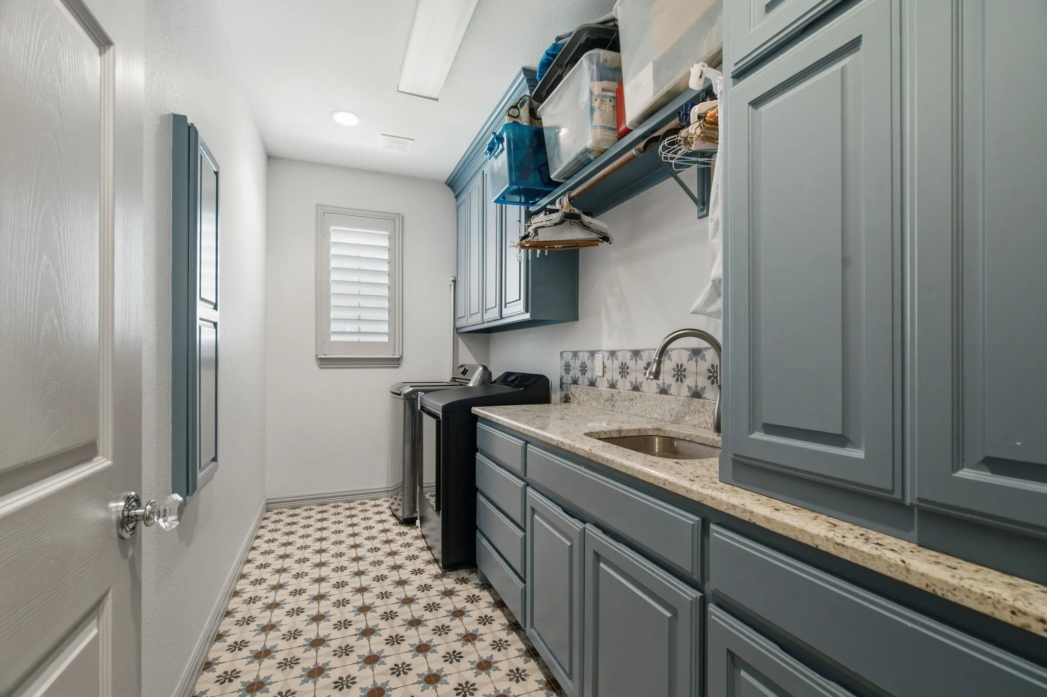 Utility room featuring patterned tile flooring, blue cabinetry, a built-in sink with a gooseneck faucet, light-toned countertops, and a window with plantation shutters