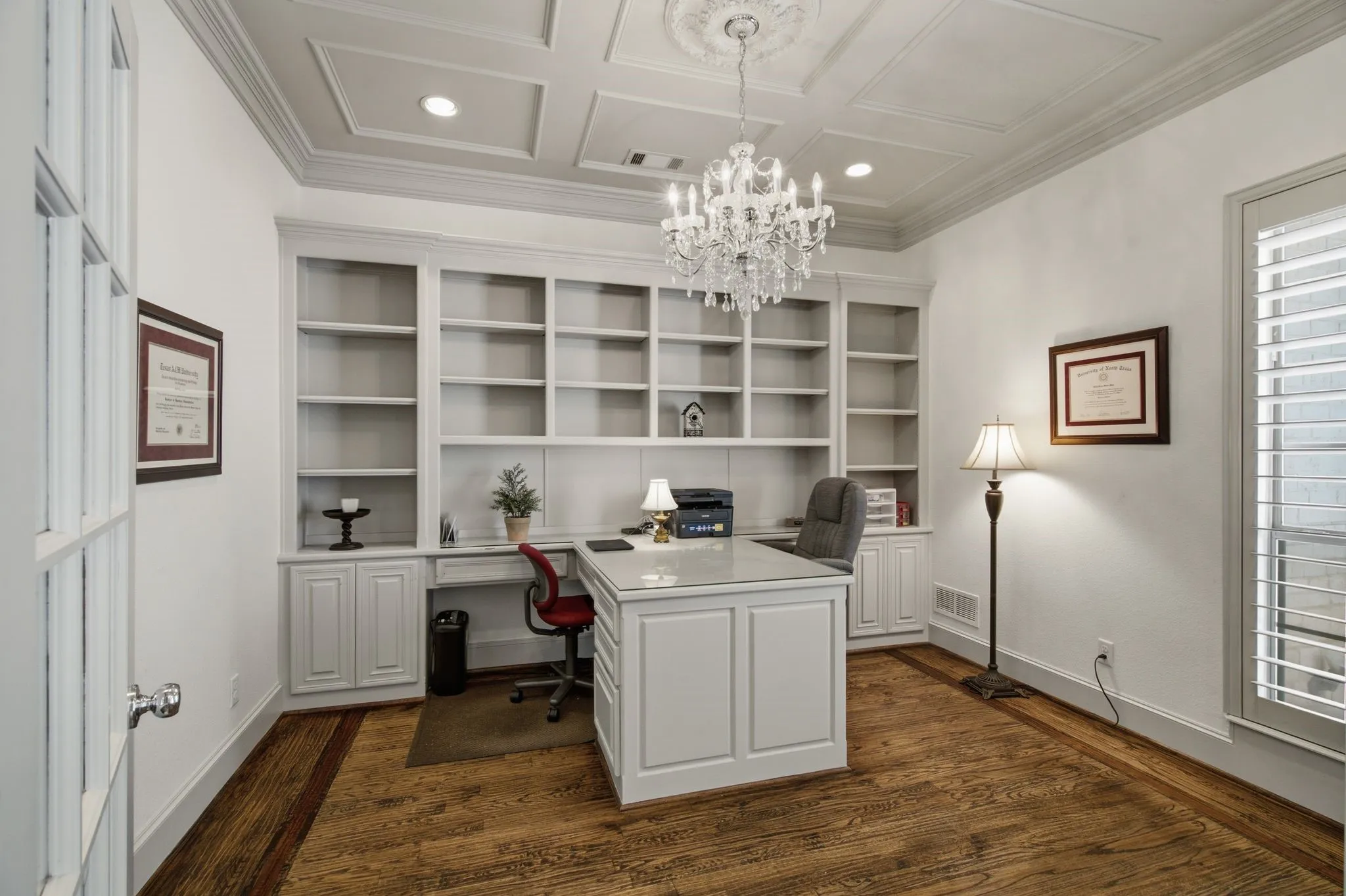 Custom built-in shelving with integrated desk space, coffered ceiling with central chandelier, wood-finish flooring, and white plantation shutters