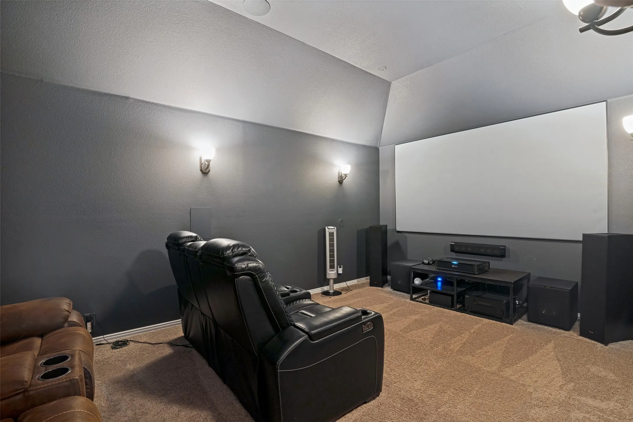 Dedicated media room featuring a large projection screen, recessed ceiling speakers, wall sconces, and sloped ceilings