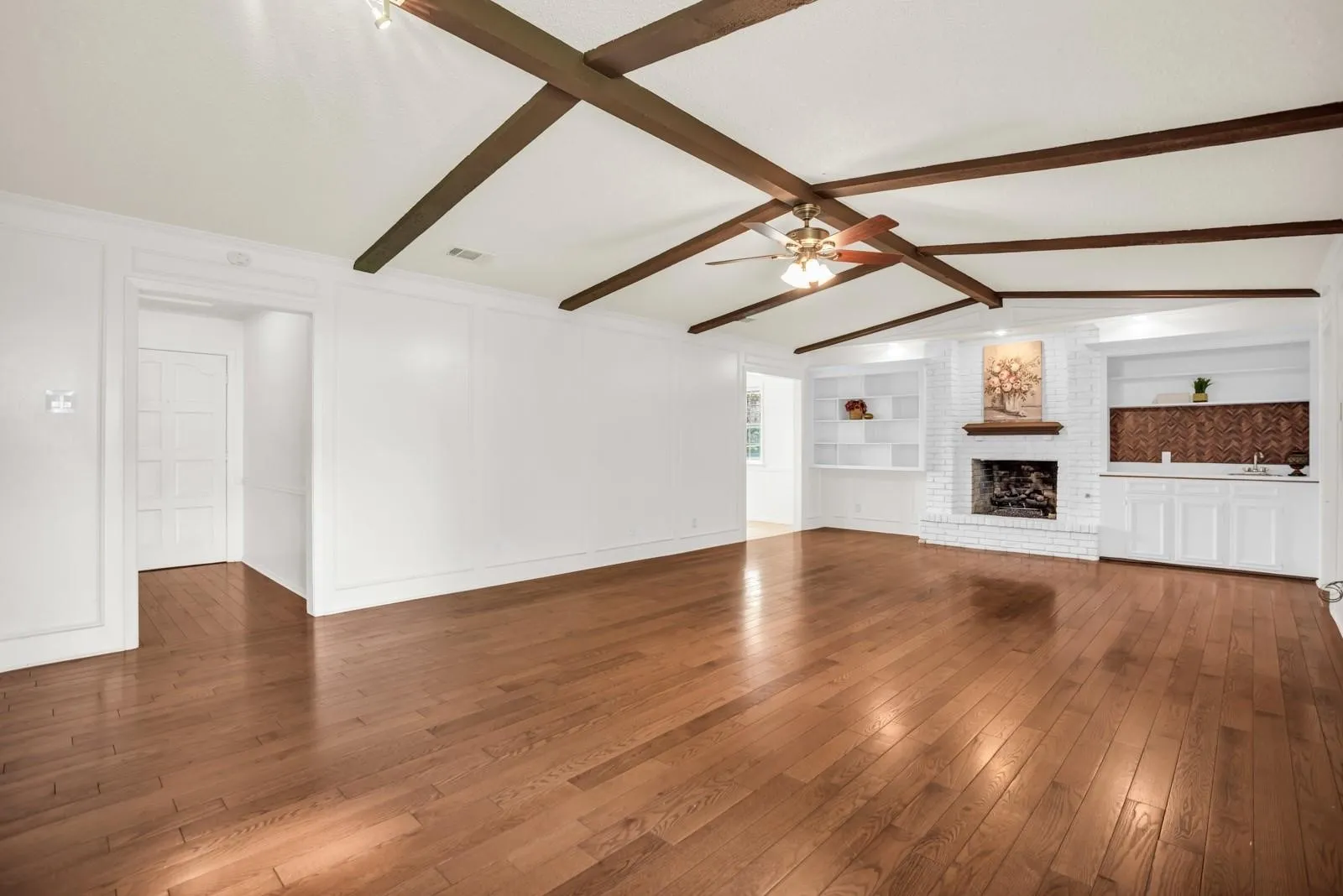 Spacious room featuring wood-finish flooring, exposed ceiling beams, and a white brick fireplace with a wood mantel