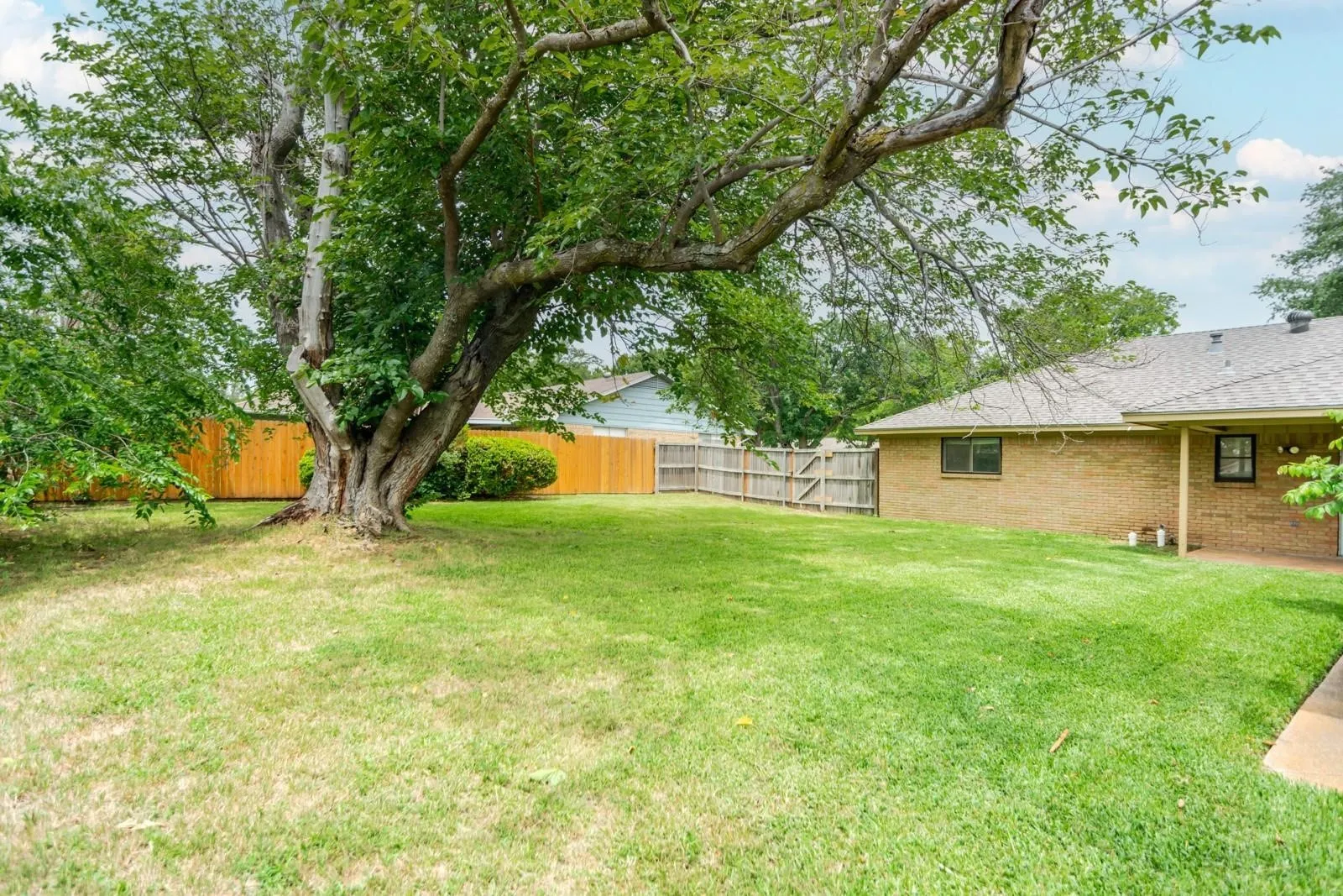 Expansive backyard featuring a mature shade tree, a wood privacy fence, and a brick exterior home with a covered patio