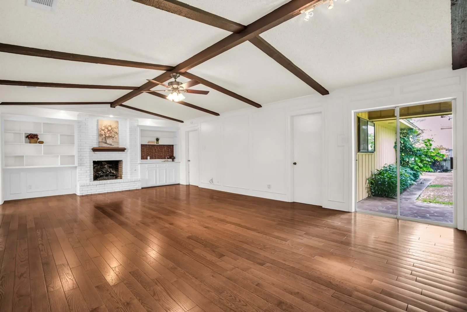 Spacious living area featuring exposed ceiling beams, wood-finish flooring, a brick fireplace with a wood mantel, built-in shelving, and sliding glass doors opening to the exterior