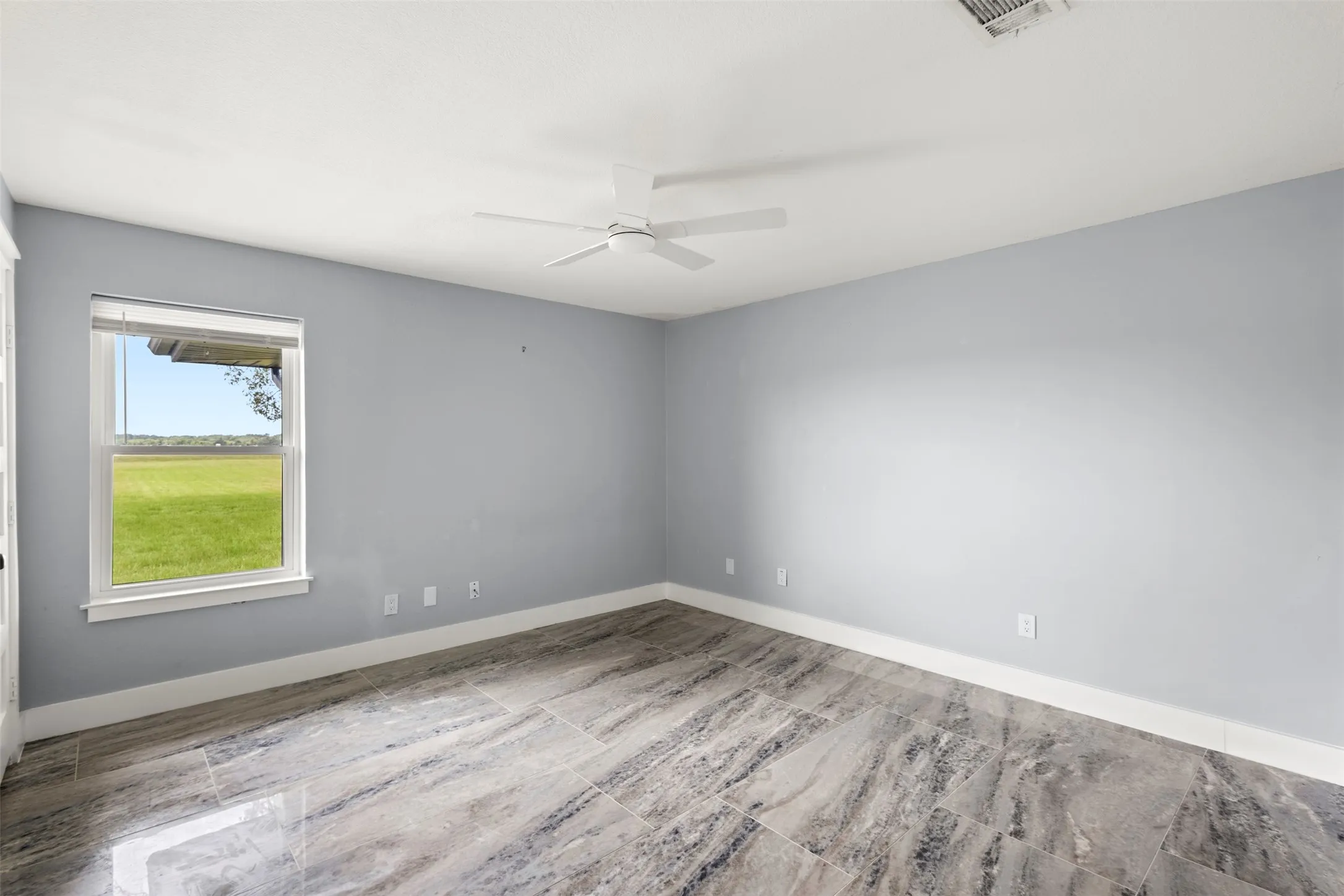 Room featuring large format tile flooring, a window with outdoor scenery, a white ceiling fan, and light blue walls with white baseboards
