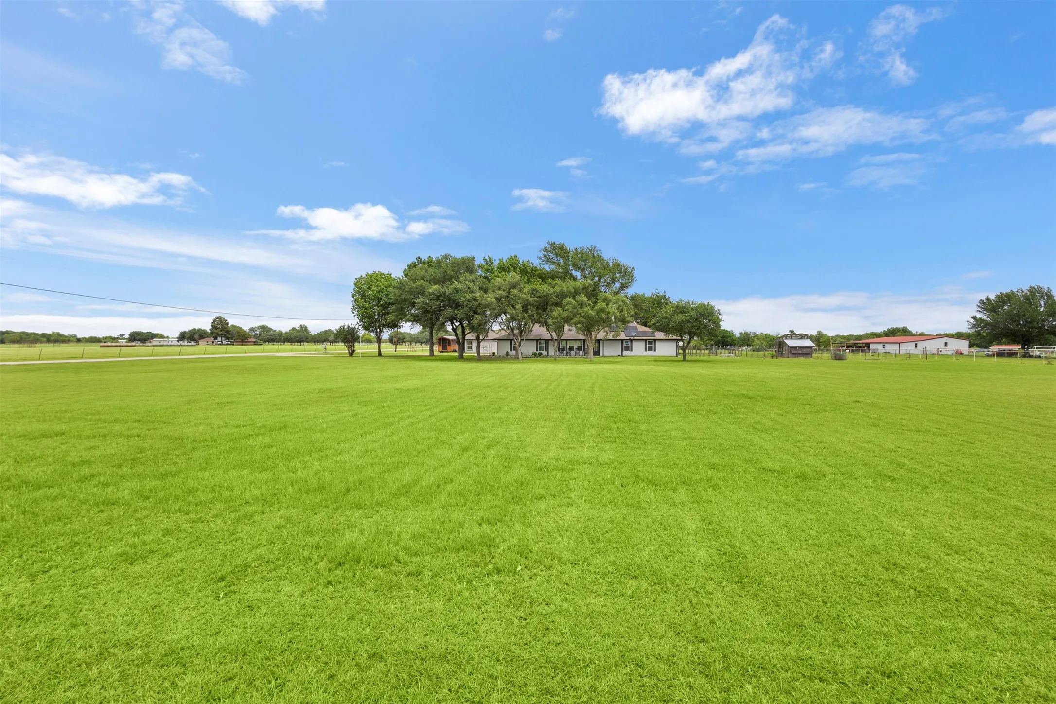 Expansive green field with a distant white residence featuring a dark roof and surrounding mature trees