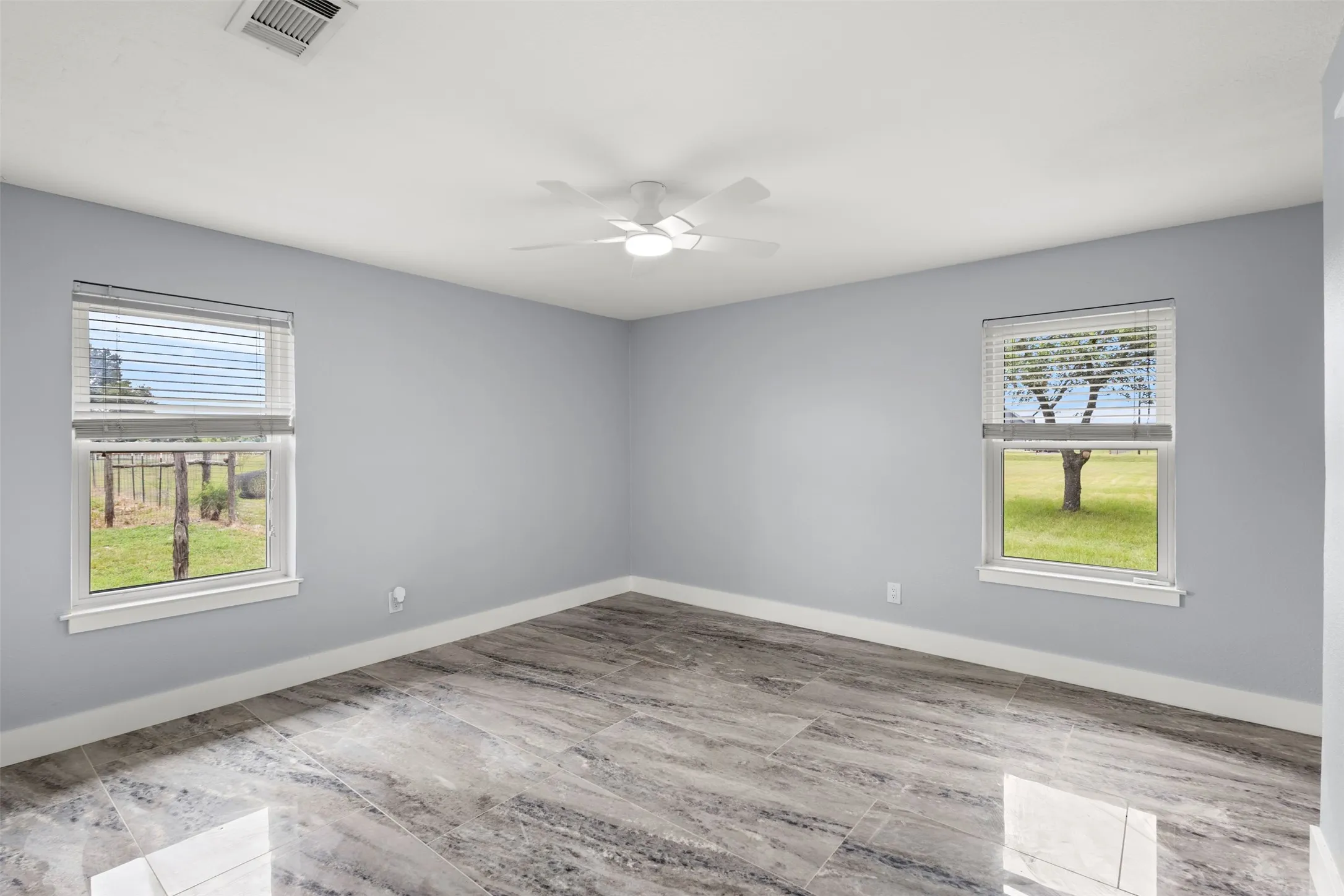 Spacious room featuring wood-finish flooring, two large windows with horizontal blinds, a ceiling fan with integrated lighting, and light-toned walls
