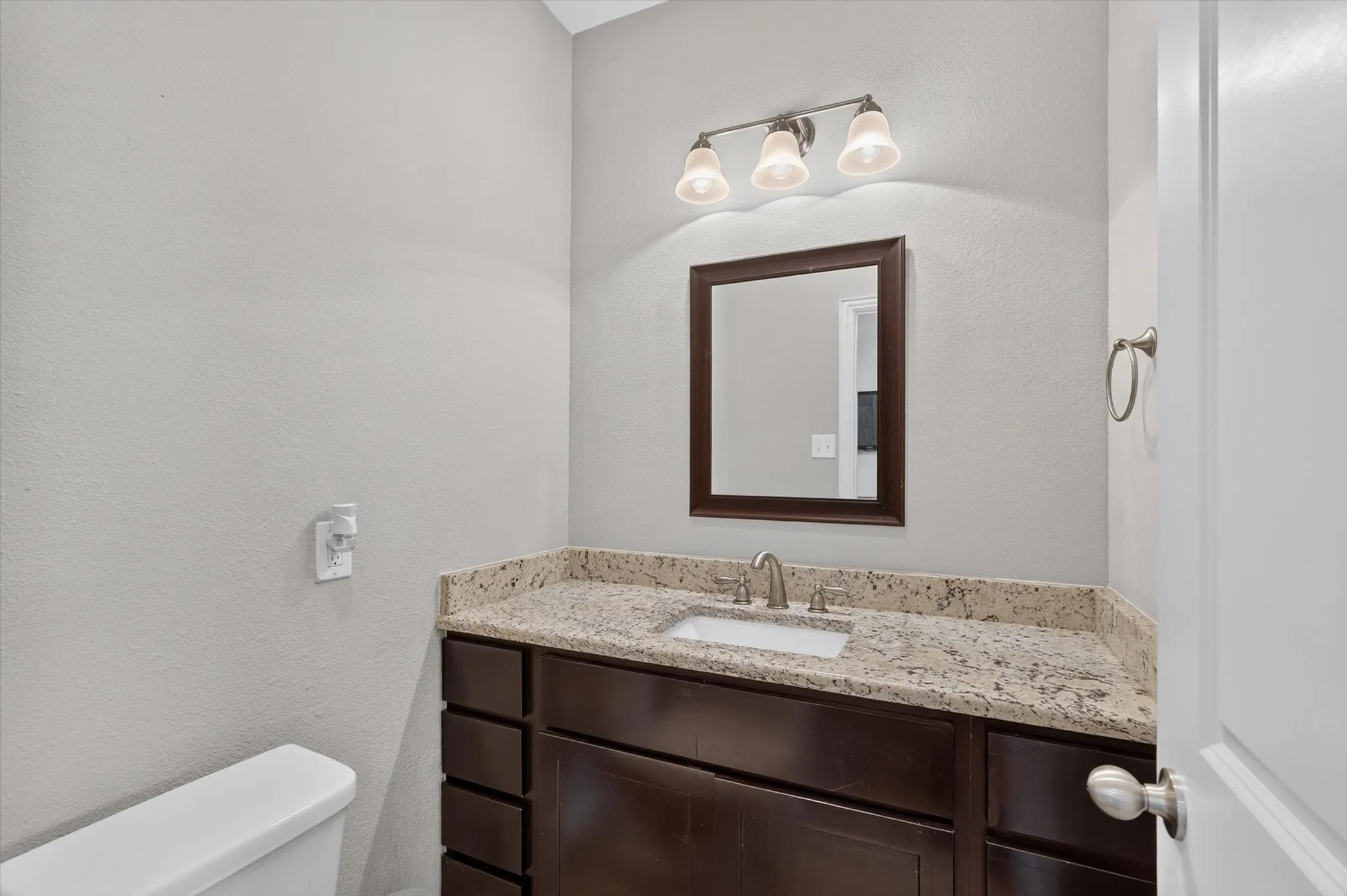 Bathroom vanity featuring a granite-style countertop, undermount sink, dark wood-finish cabinetry, and a framed mirror with a three-light fixture