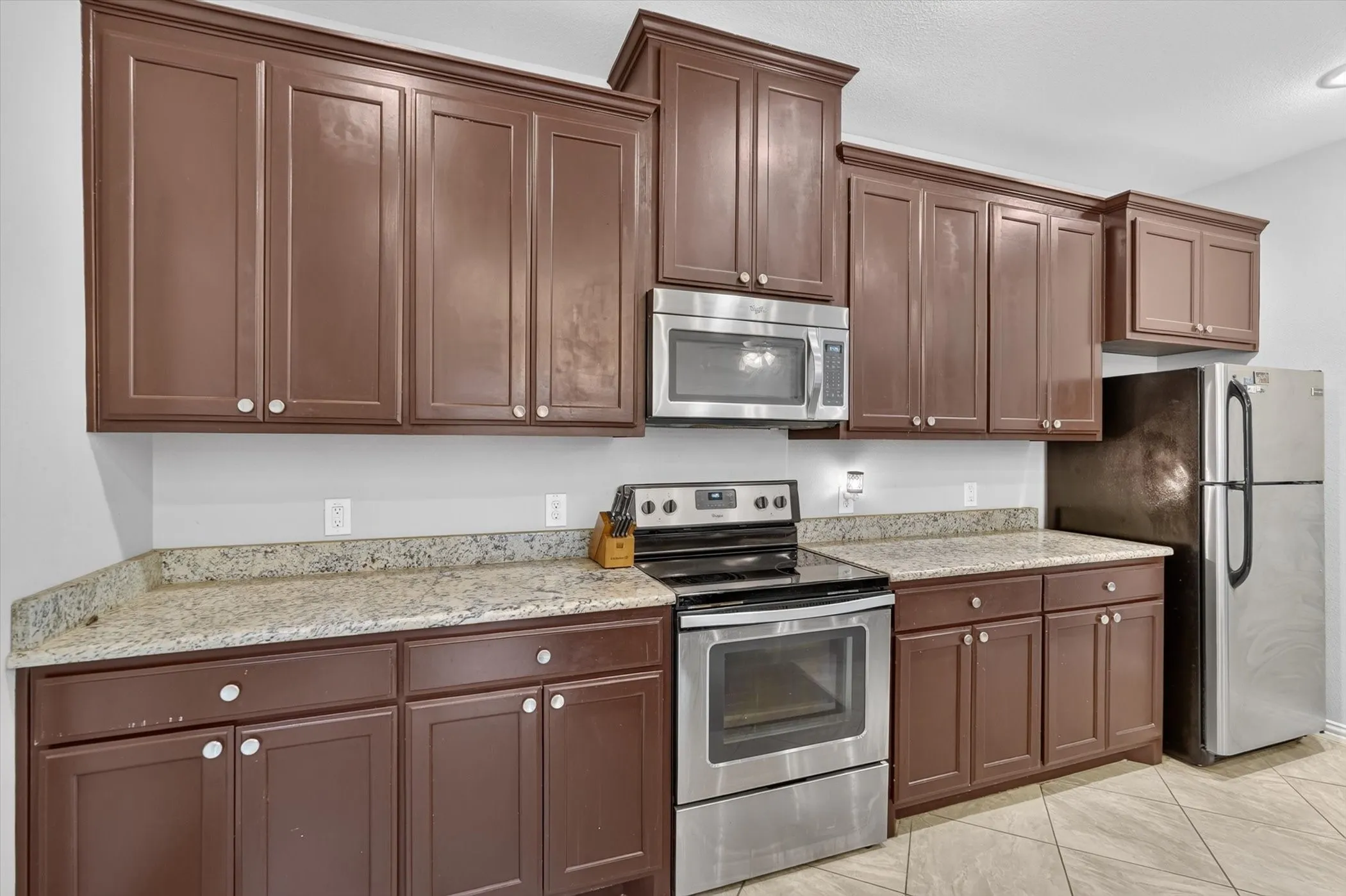 Kitchen featuring brown cabinetry with white knobs, light granite-style countertops, stainless steel appliances, and tiled flooring