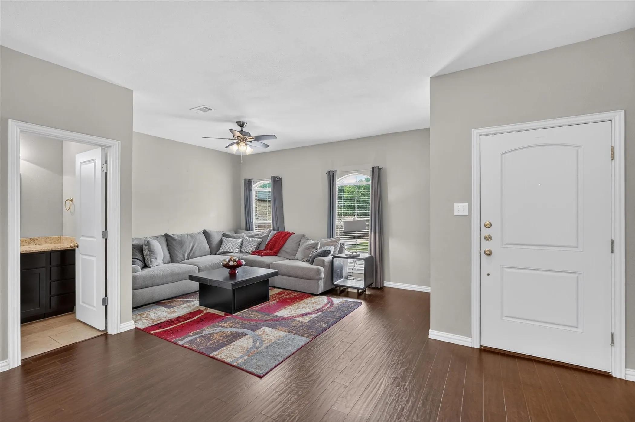 Living area featuring wood-finish flooring, light gray wall paint, two windows with blinds, and a ceiling fan with integrated lighting