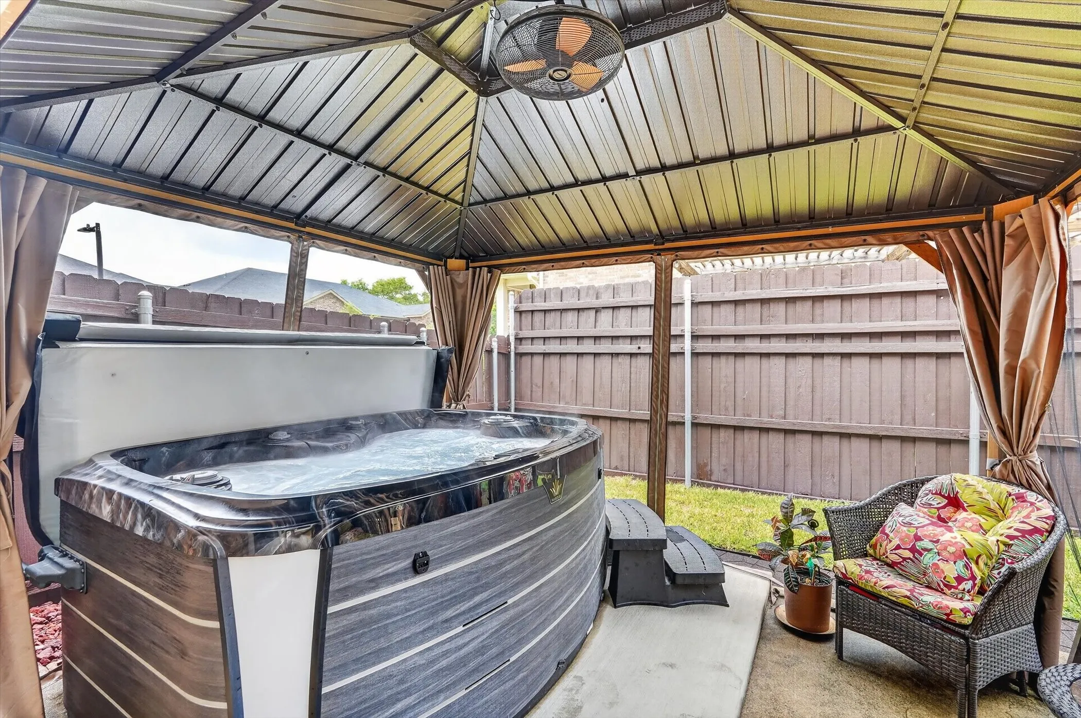 Outdoor spa with a dark wood-finish exterior, enclosed within a metal gazebo featuring a ceiling fan and privacy curtains