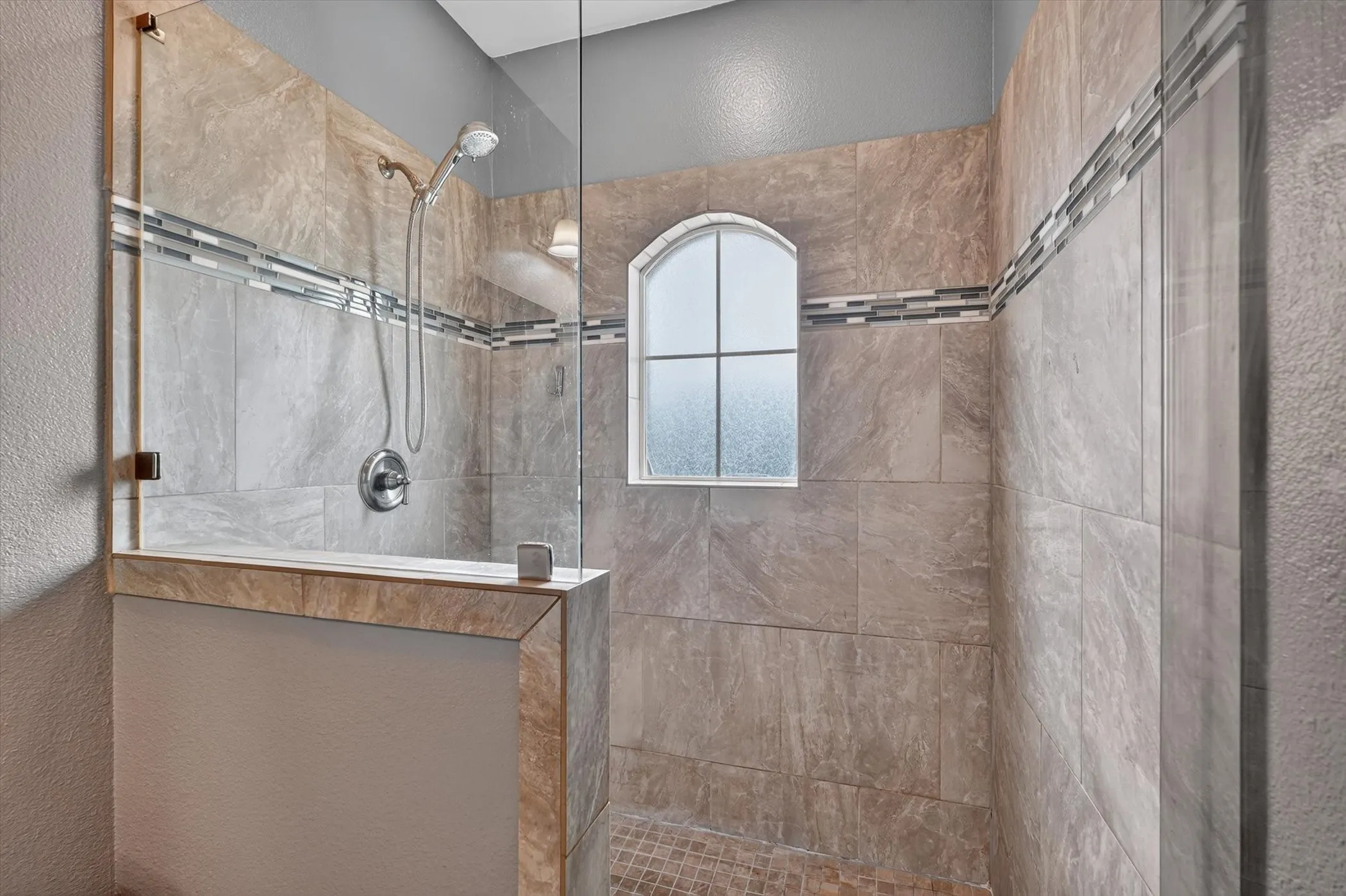 Glass-enclosed shower featuring large format wall tiles, decorative accent banding, an arched window, a tiled shower pan, and a handheld showerhead with a wall-mounted control