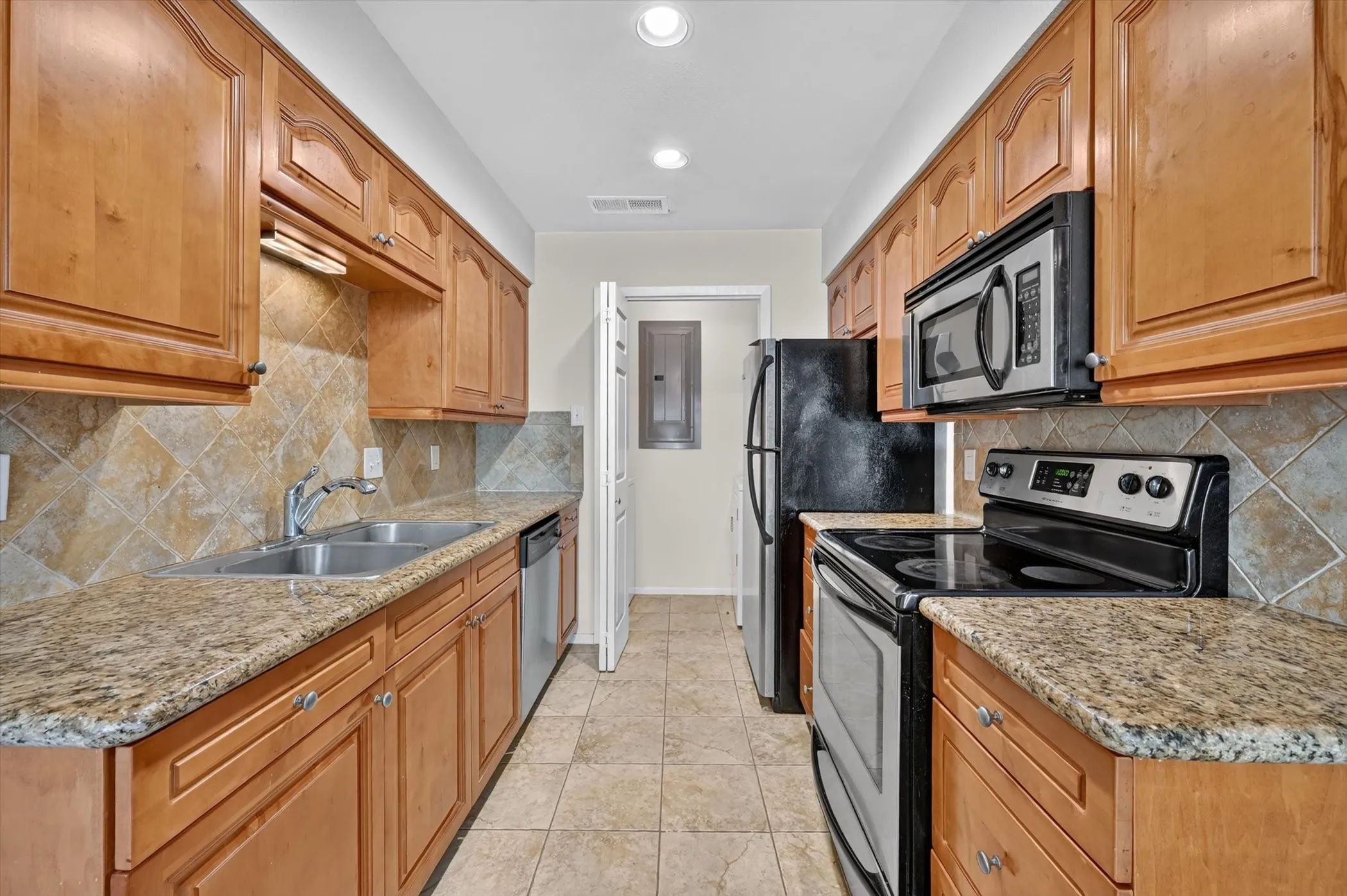 Galley kitchen featuring light wood cabinetry, stone countertops, a stainless steel double basin sink, and tile flooring