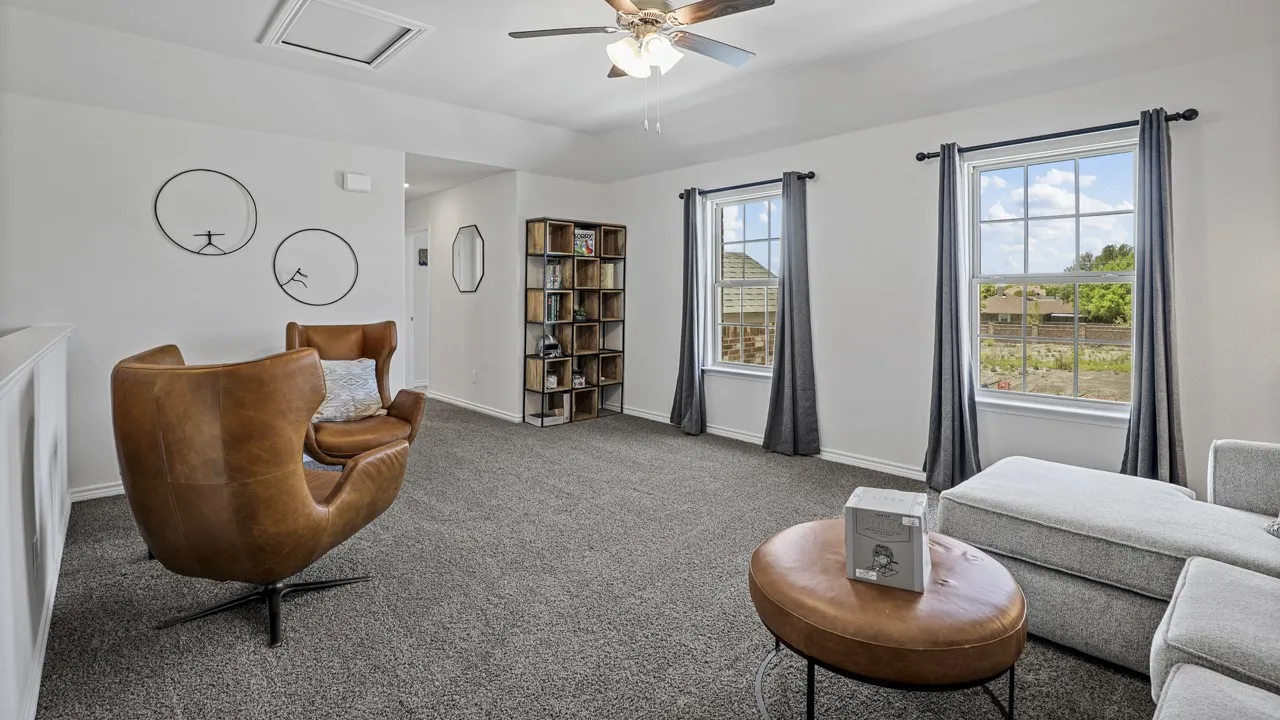 Carpeted upstairs bonus room featuring a ceiling fan with light fixture, two double-hung windows with grid patterns, and a built-in shelving unit