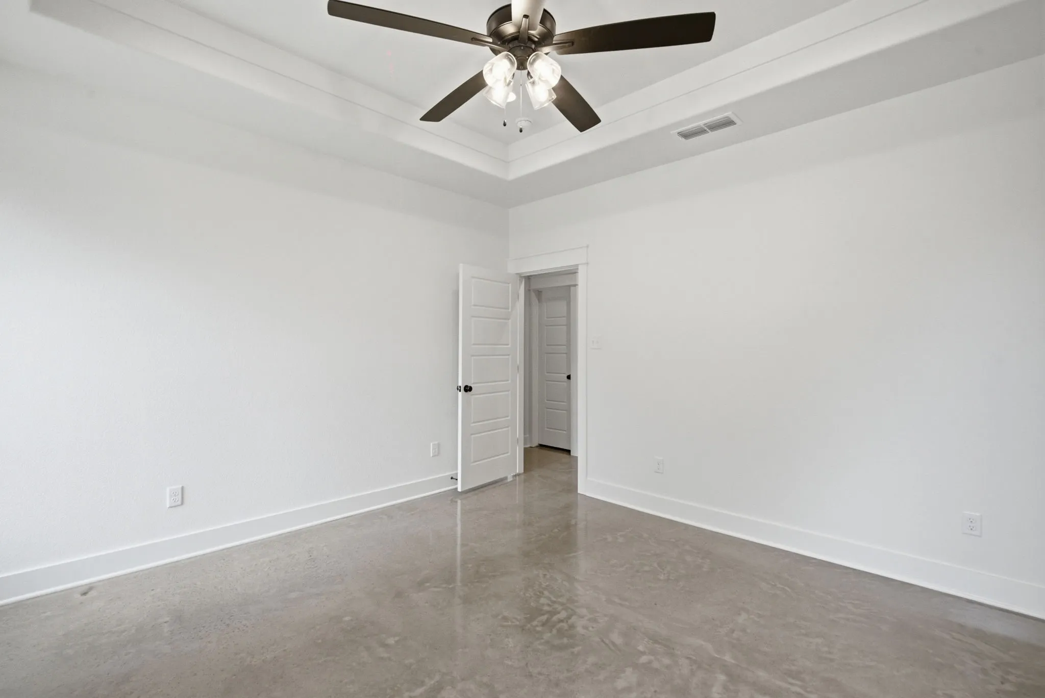 Spacious room featuring tray ceilings, a dark-toned ceiling fan with integrated lighting, polished concrete flooring, white baseboards, and a paneled interior door