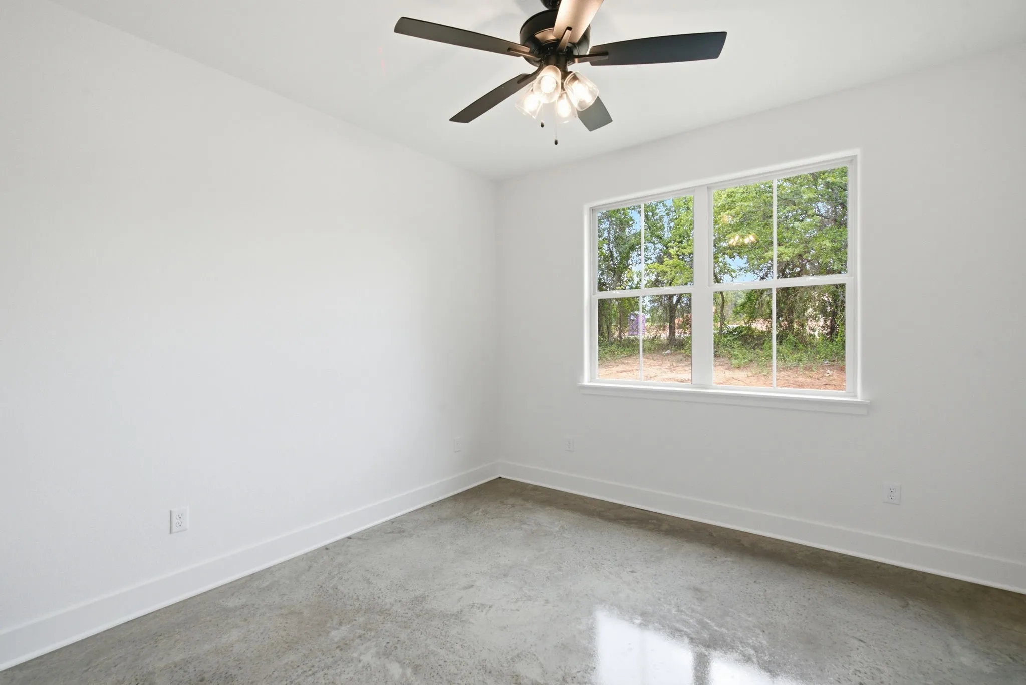 Spacious room featuring a large window, a ceiling fan with integrated lighting, concrete flooring, and crisp white baseboards