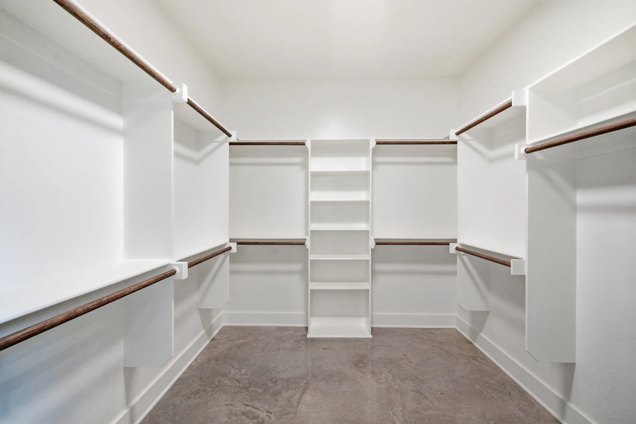 Spacious walk-in closet featuring built-in shelving, multiple hanging rods, and a central storage tower, all finished in white with dark-stained wood rods