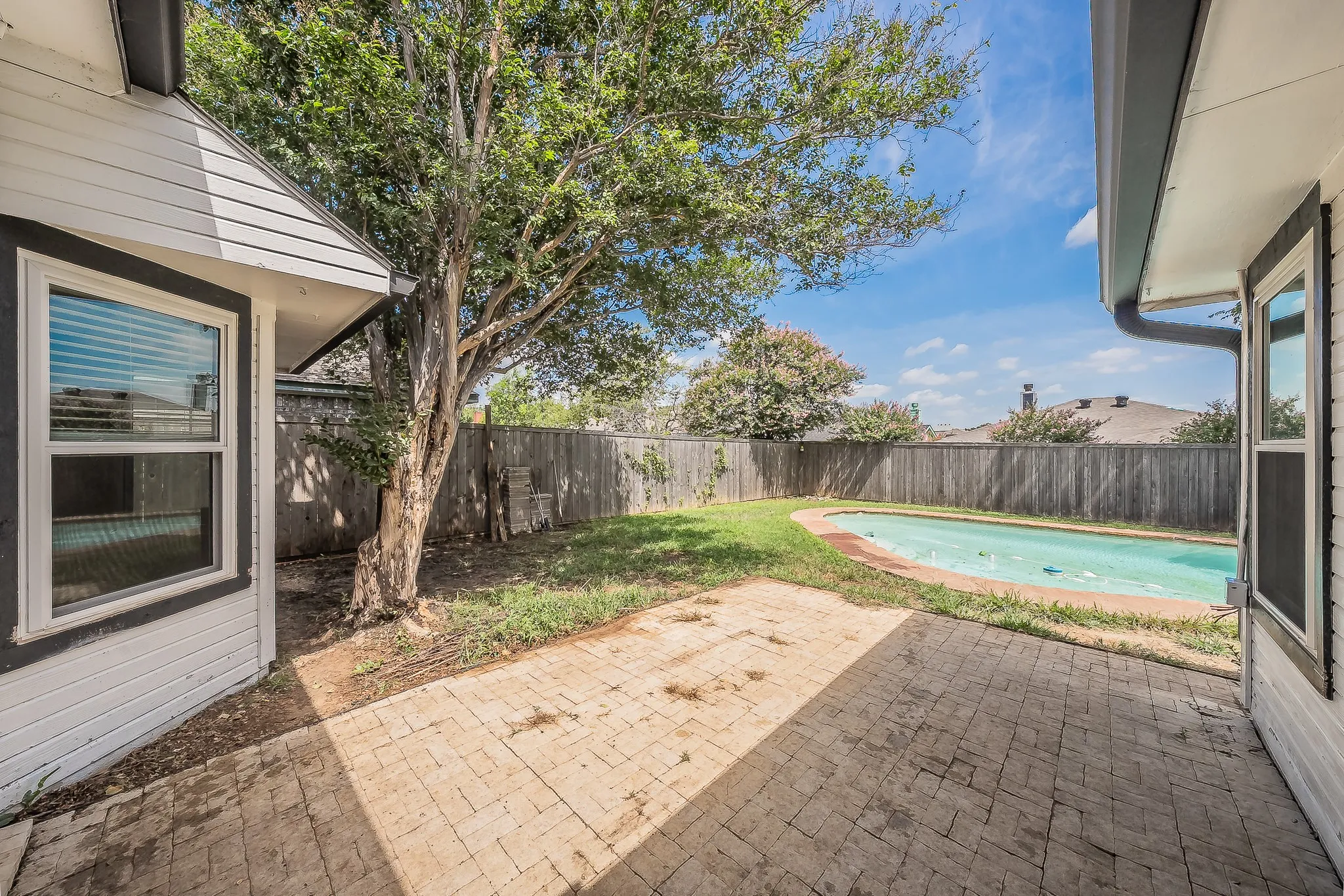 Brick paver patio with a curved swimming pool, surrounded by a wooden privacy fence, mature tree, and exterior siding with dark trim