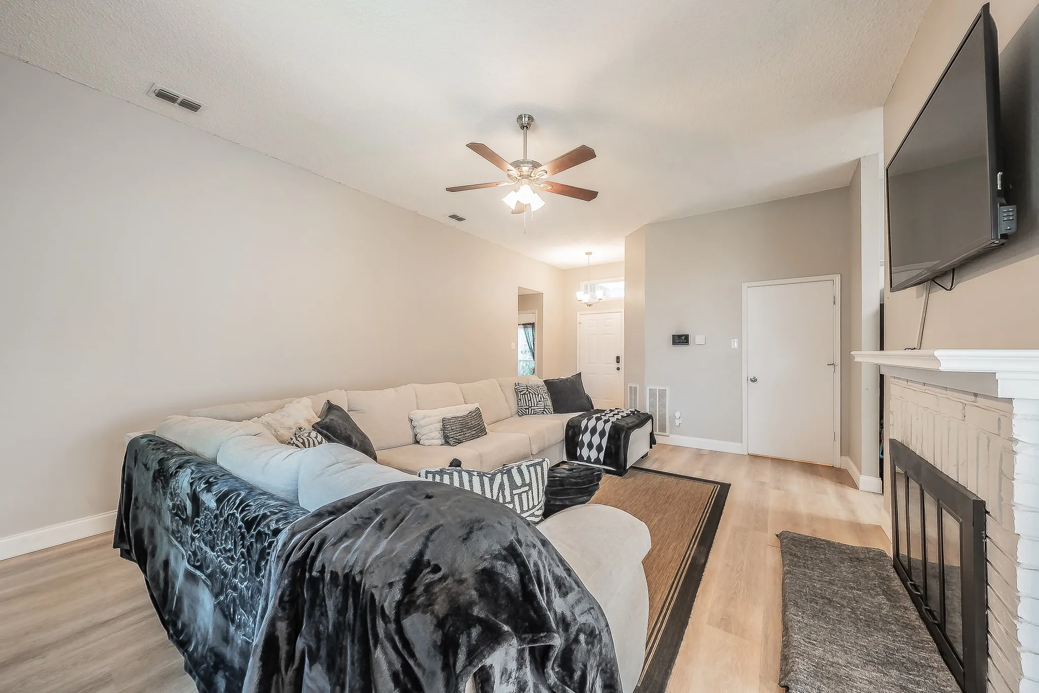 Spacious living area featuring light wood-finish flooring, a painted brick fireplace with a white mantel, and a ceiling fan