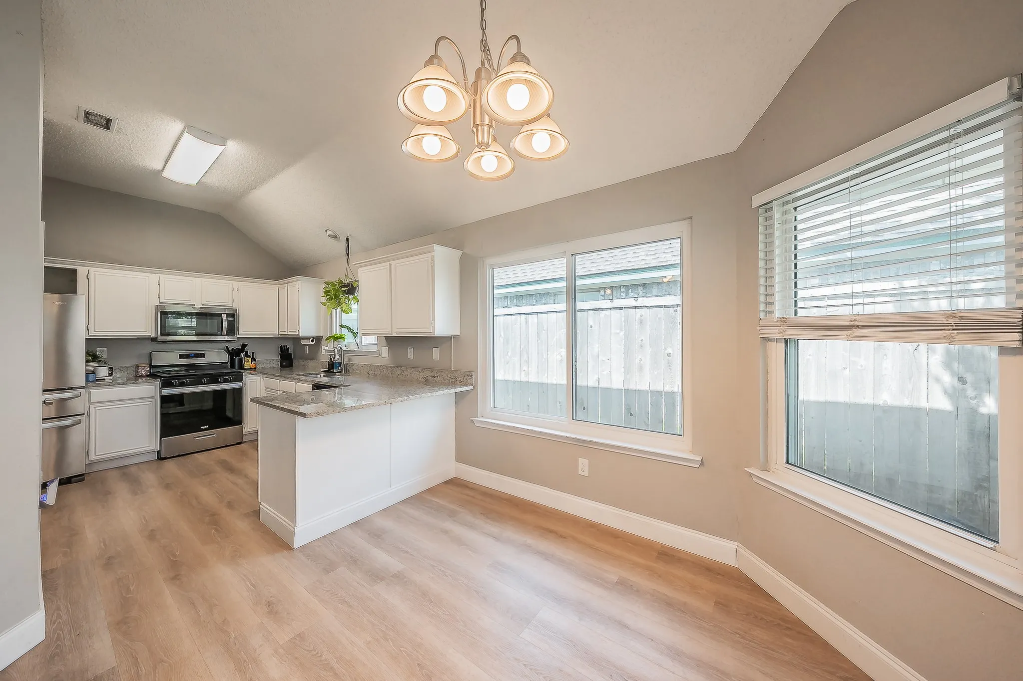 Open-concept kitchen and dining area featuring vaulted ceilings, light wood-finish flooring, white cabinetry, stainless steel appliances, and granite-style countertops