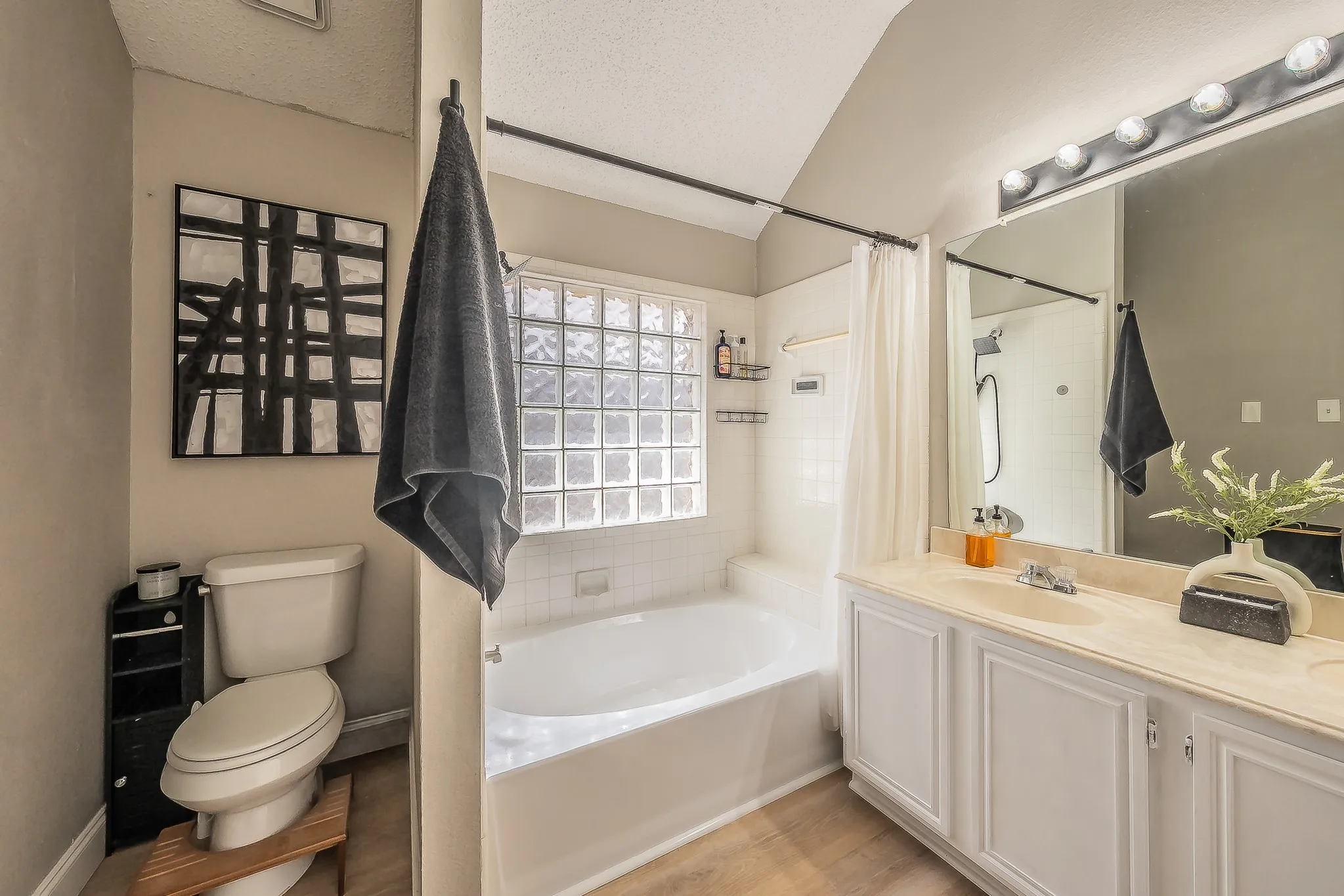 Bathroom featuring a bathtub with glass block window, a vanity with dual sinks and light-up mirror, wood-finish flooring, and a shower with white tile surround