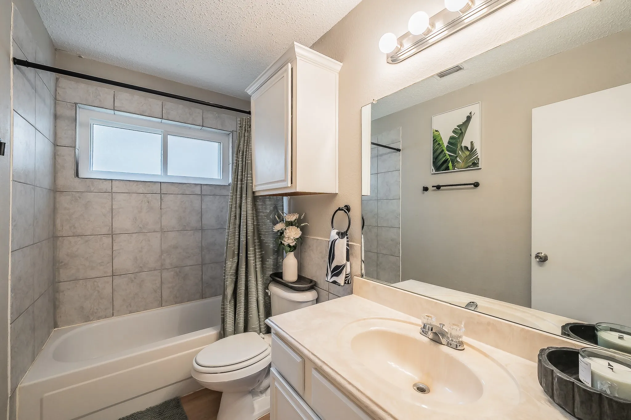 Bathroom featuring a single vanity with an integrated sink, a large mirror, overhead vanity lighting, a built-in cabinet, and a tiled shower-over-tub with a frosted window