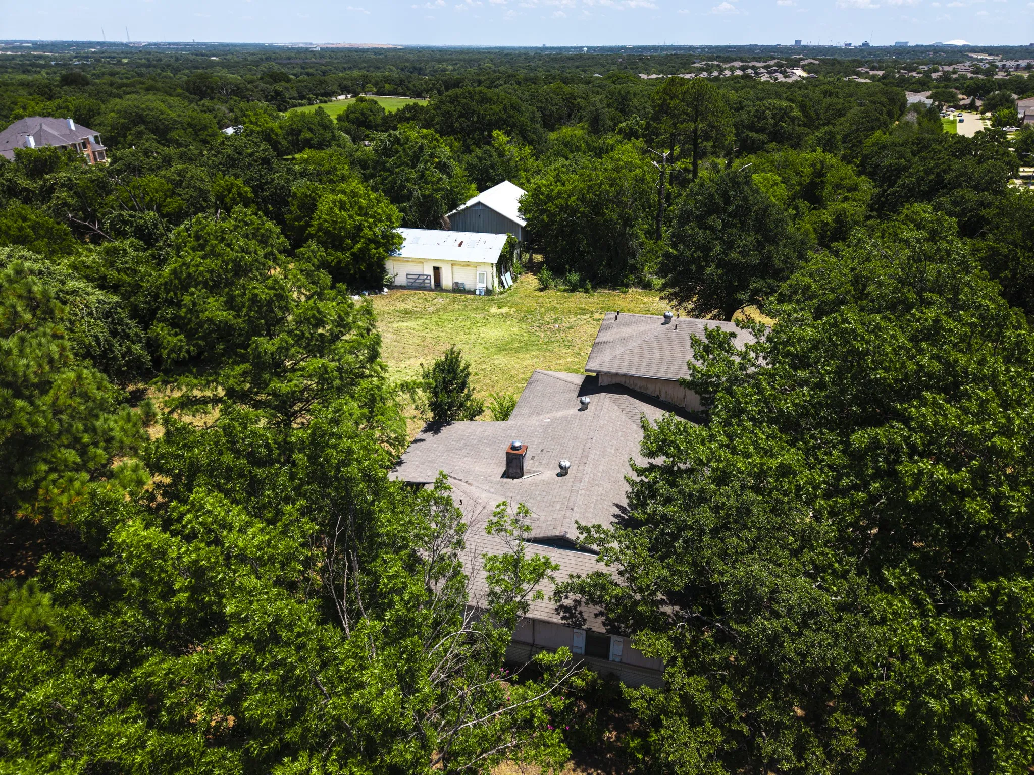 Expansive treed lot featuring a residential structure with a shingled roof and a detached outbuilding with a metal roof