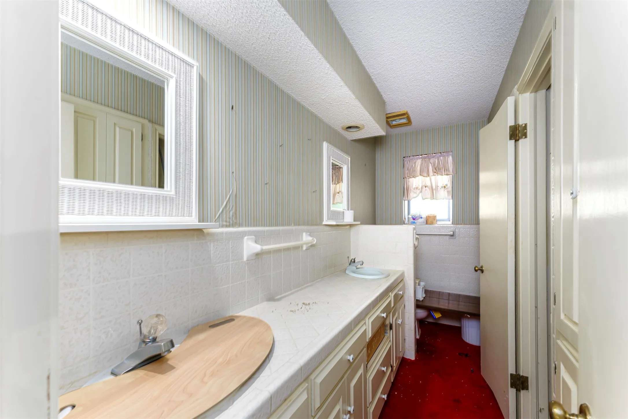 Bathroom featuring a vanity with a white countertop and base cabinetry, a wall-mounted mirror, light-toned striped wallpaper, and a window with a valance