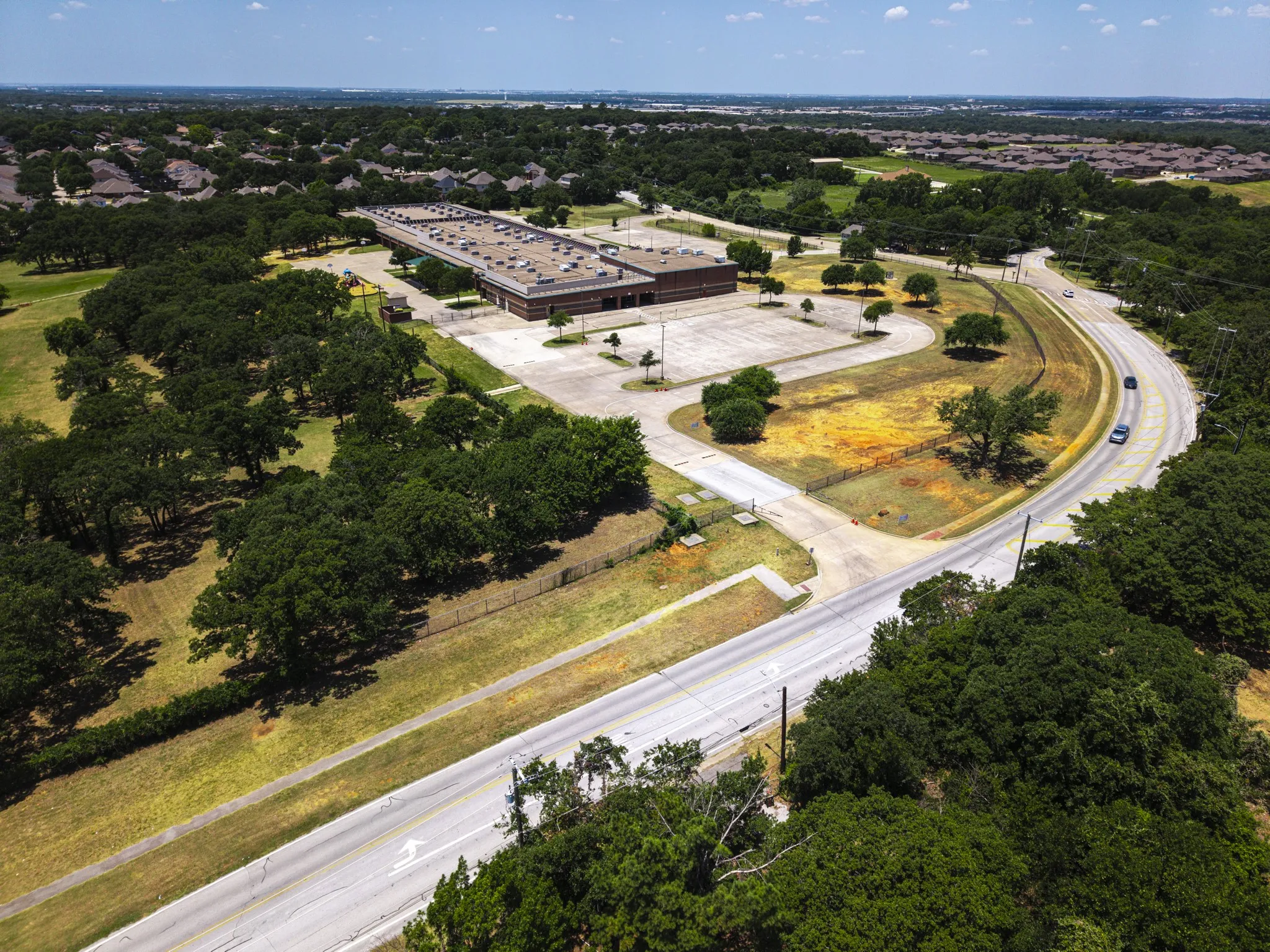 Aerial perspective showcasing a large institutional building with an expansive paved lot, surrounded by abundant mature trees and verdant green spaces