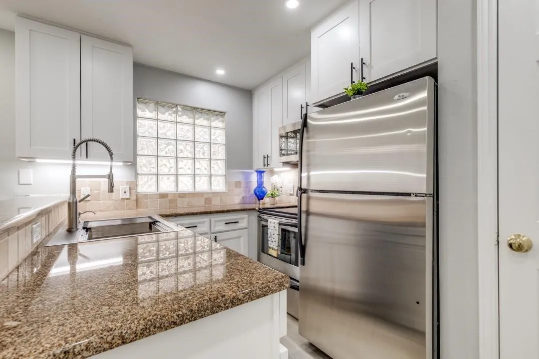 Modern kitchen featuring white cabinetry, granite-finish countertops, stainless steel appliances, a tile backsplash, and a glass block window