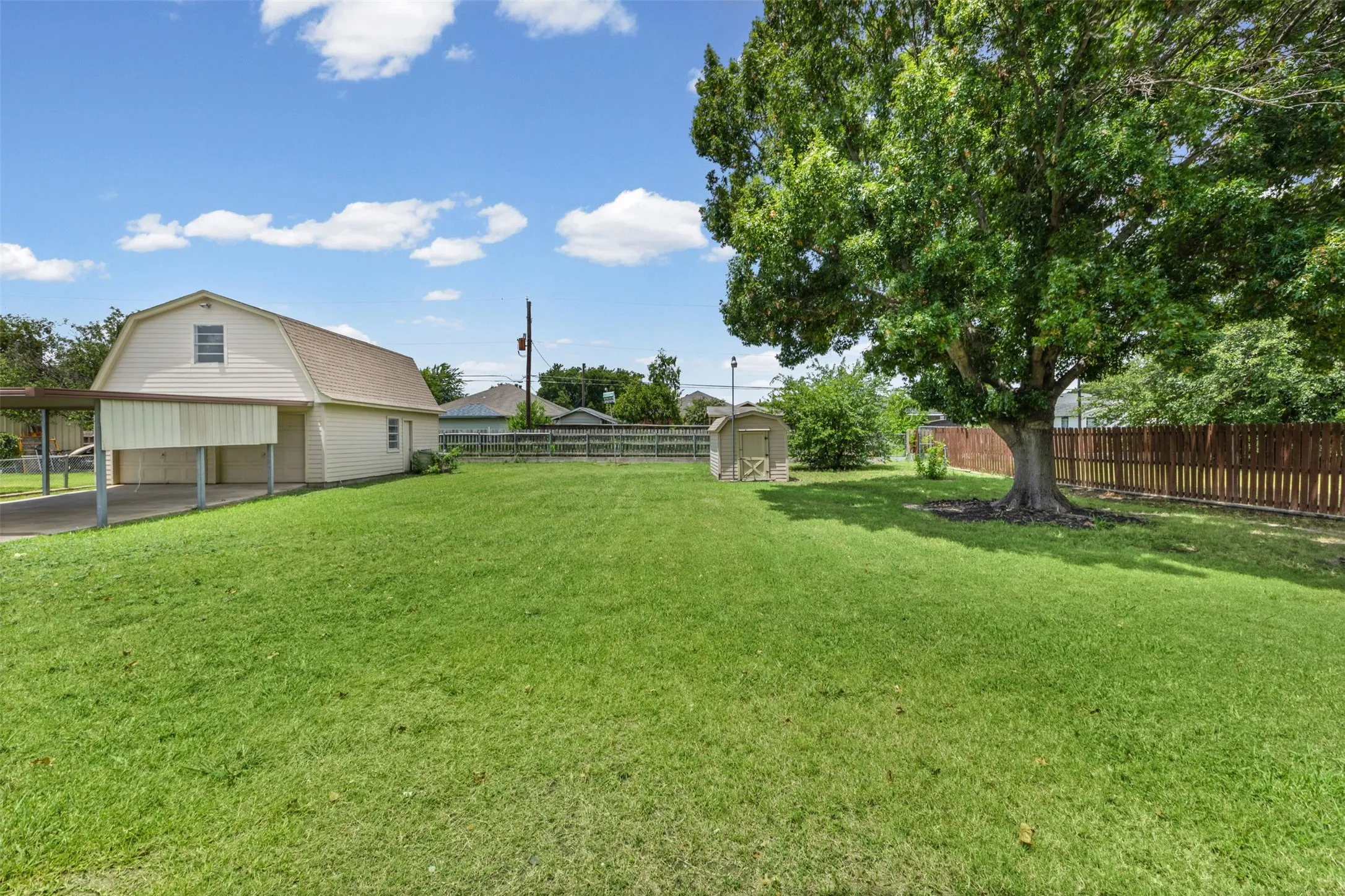 Expansive green lawn featuring a detached accessory building, a large mature tree, a wooden privacy fence, and an additional storage shed