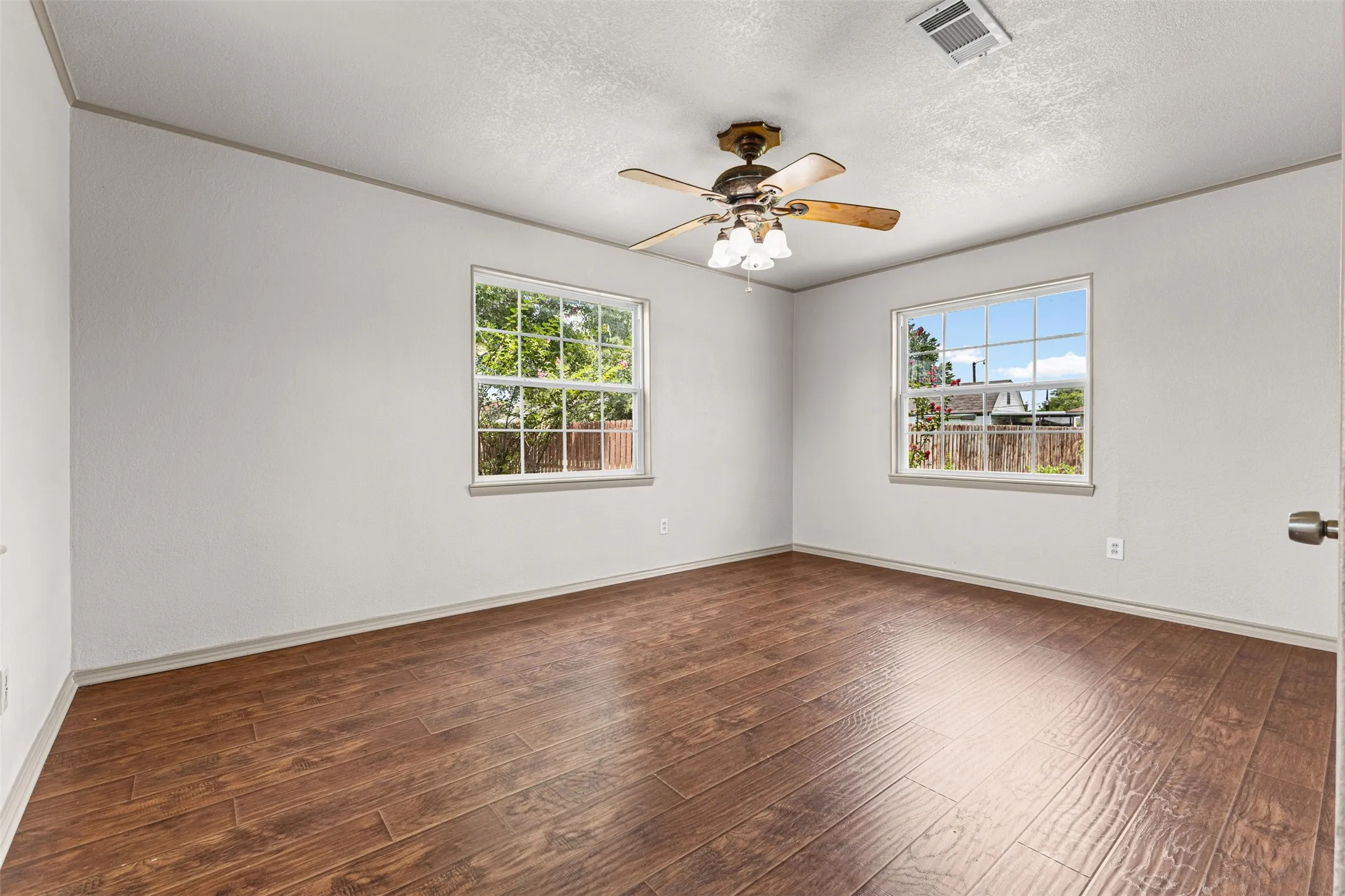 Spacious room featuring wood-finish flooring, two large windows, a ceiling fan with integrated lighting, and neutral wall tones
