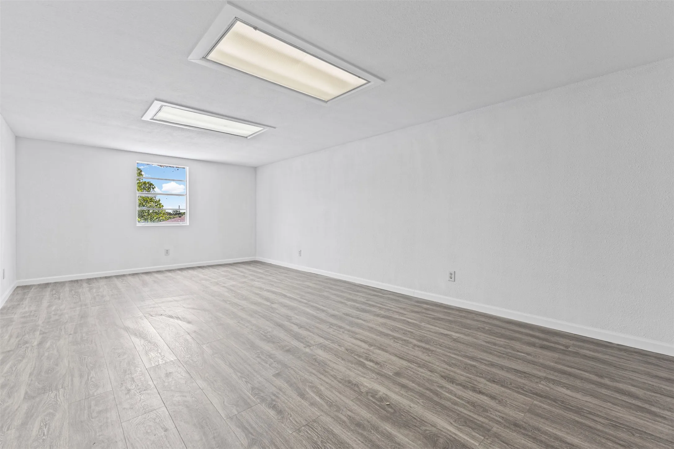Spacious room featuring wood-finish flooring, white walls, and a single window offering exterior views