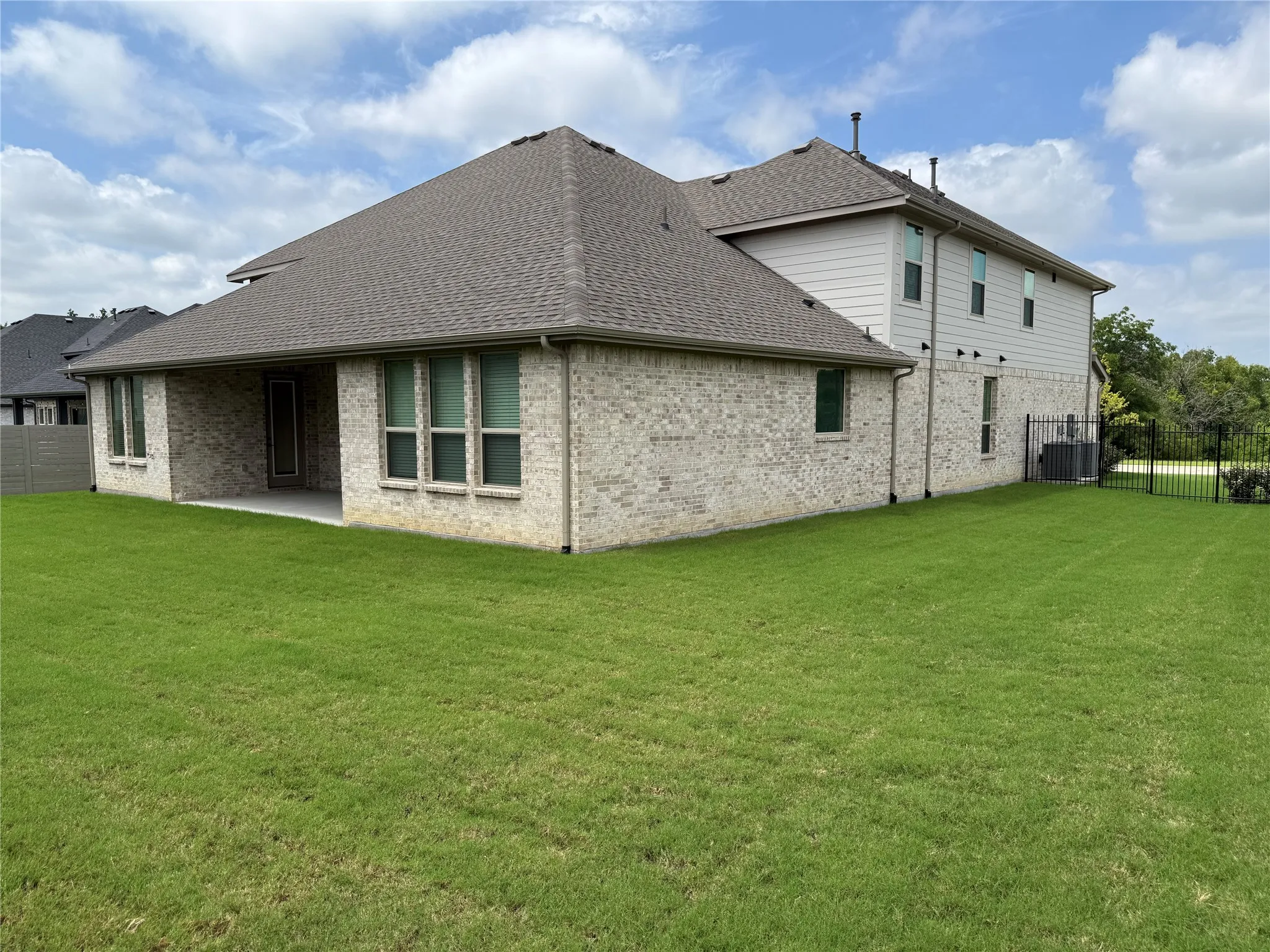Spacious rear yard with a covered patio, brick and siding exterior, multiple windows, and a black wrought iron fence