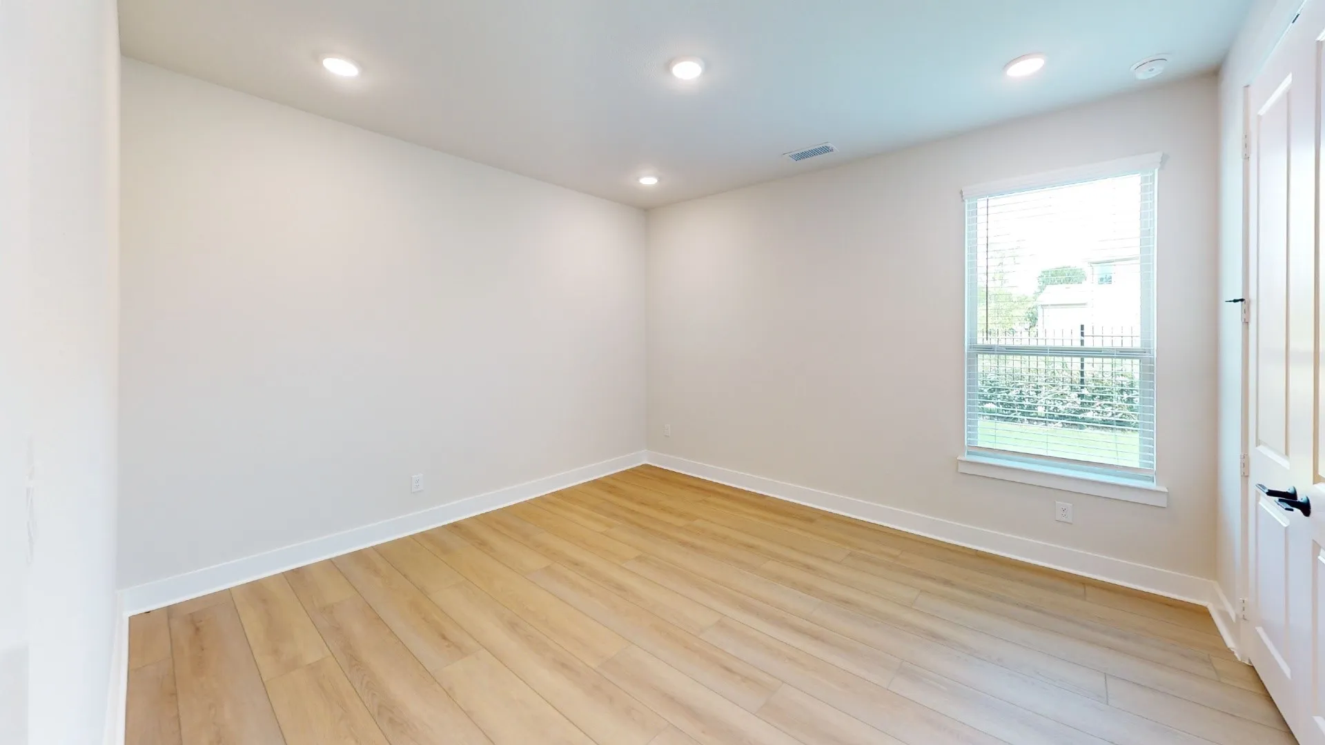 Bright interior space featuring light wood-finish flooring, recessed lighting, a single window with blinds, and a white panel door with dark hardware