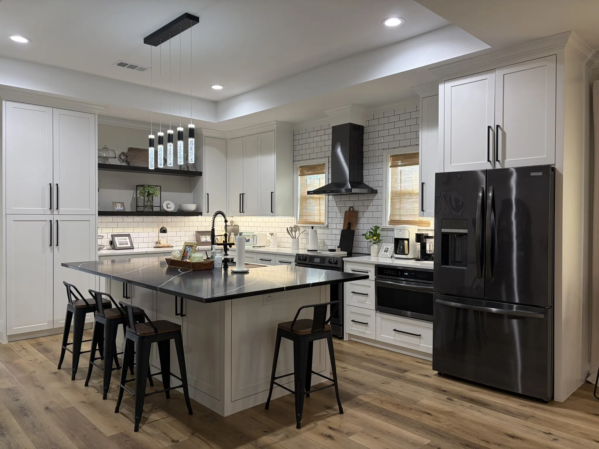 Spacious kitchen featuring an expansive center island with seating, dark stone-finish countertops, subway tile backsplash, light-toned cabinetry, and wood-finish flooring