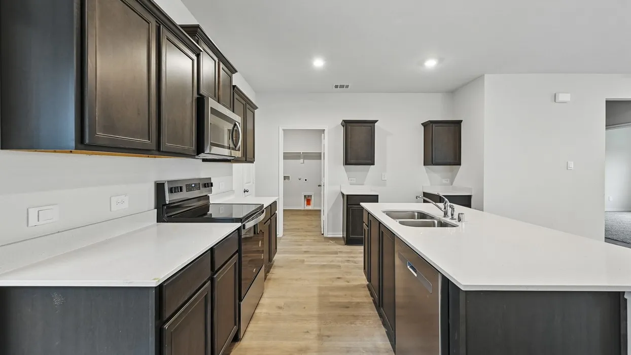 Open-concept kitchen featuring an expansive island with a double basin sink, dark wood-finish cabinetry, stainless steel appliances, light countertops, and wood-finish flooring