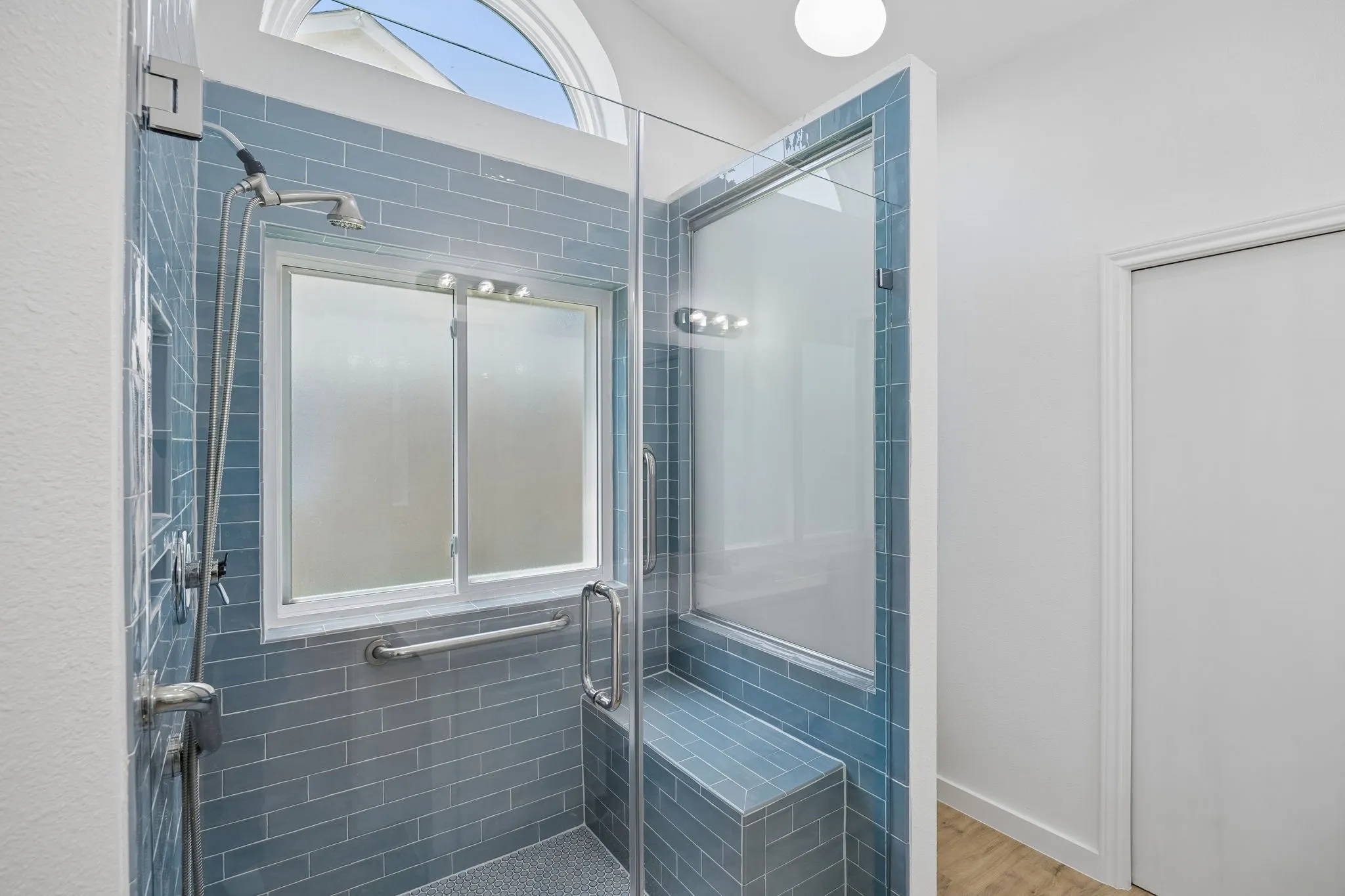 The beautifully remodeled primary bath showcases a spa-inspired walk-in shower with designer tile, frameless glass enclosure, built-in bench seating, and elegant natural light from the arched window above.