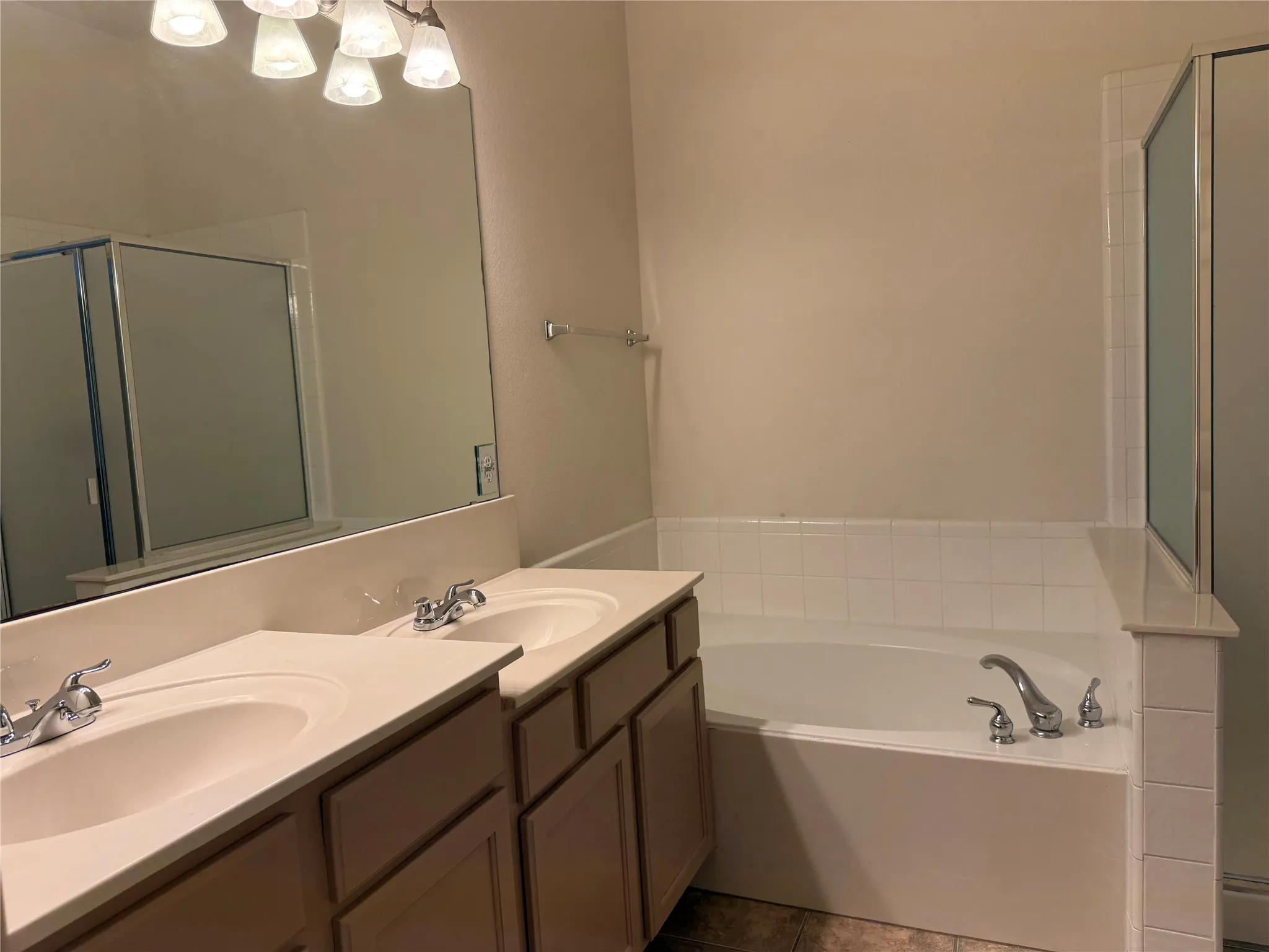 Bathroom featuring a dual vanity with integrated sinks, a large mirror, a four-light vanity fixture, a built-in garden tub with tile surround, and a glass-enclosed shower