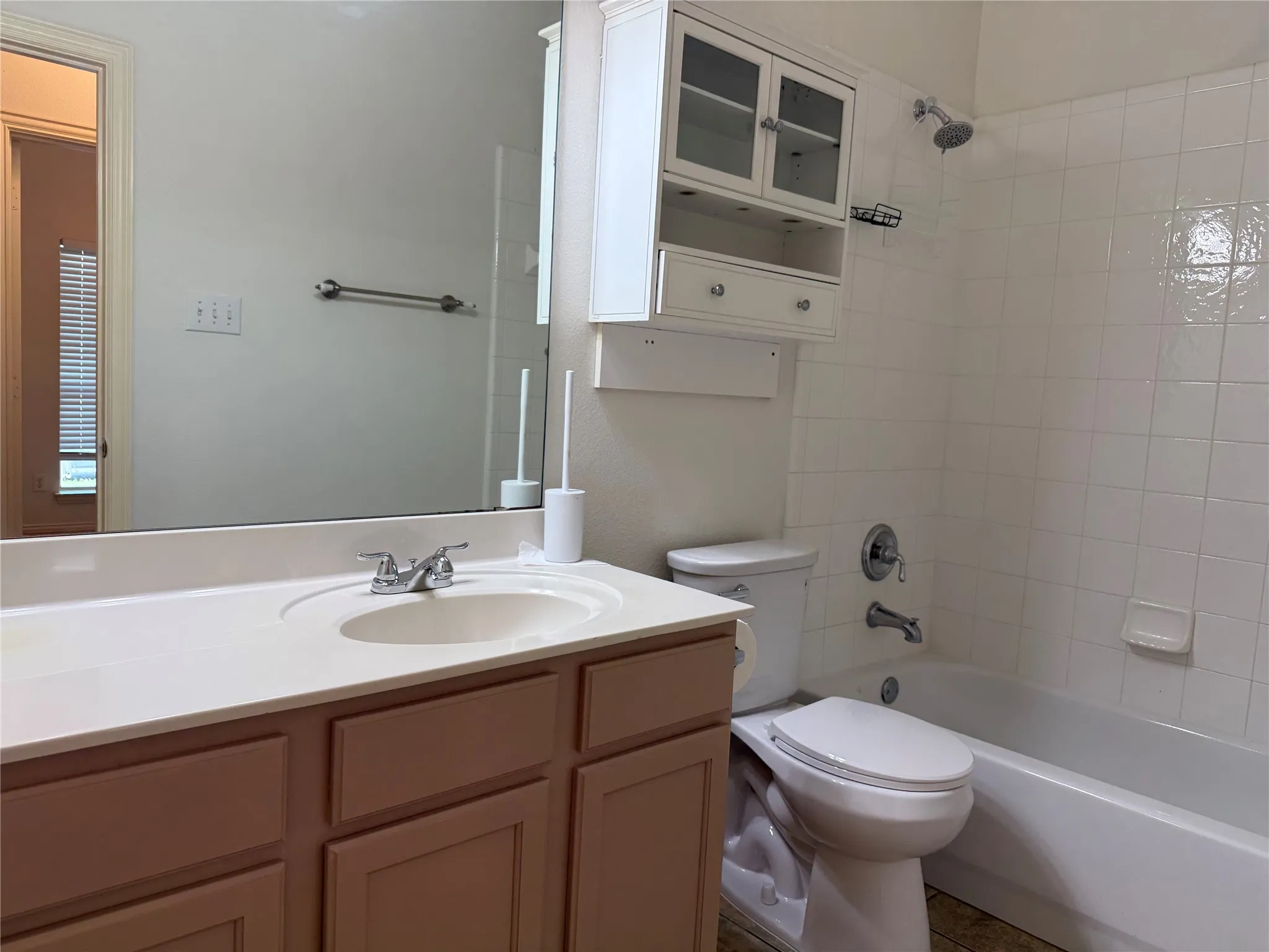 Bathroom featuring a vanity with a white countertop and integrated sink, a large wall-mounted mirror, a tub/shower combination with white tile surround, and a wall-mounted storage cabinet with glass-front doors
