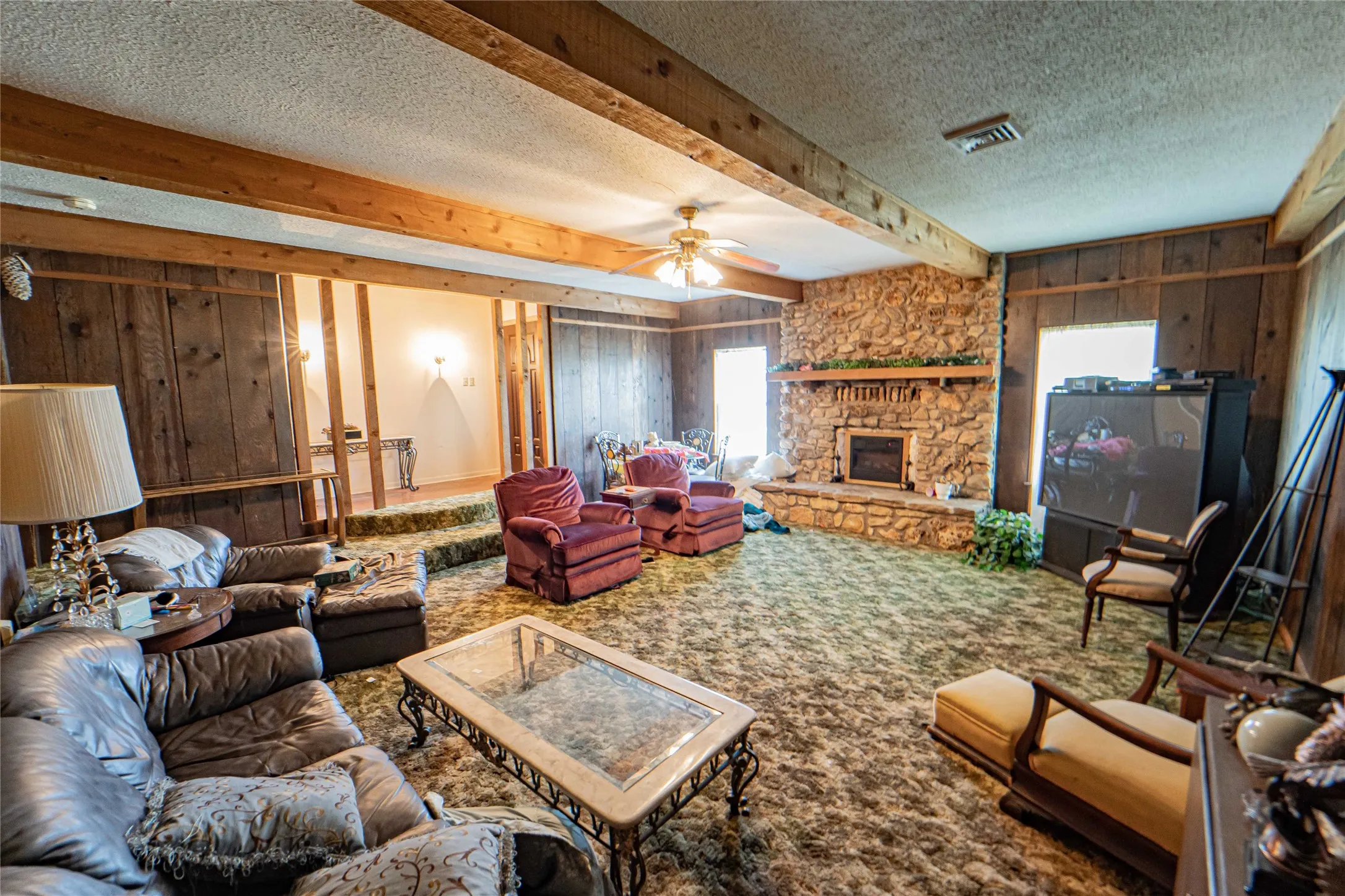 Expansive living area featuring a fireplace with hearth, wood paneling, exposed wood ceiling beams, and a ceiling fan