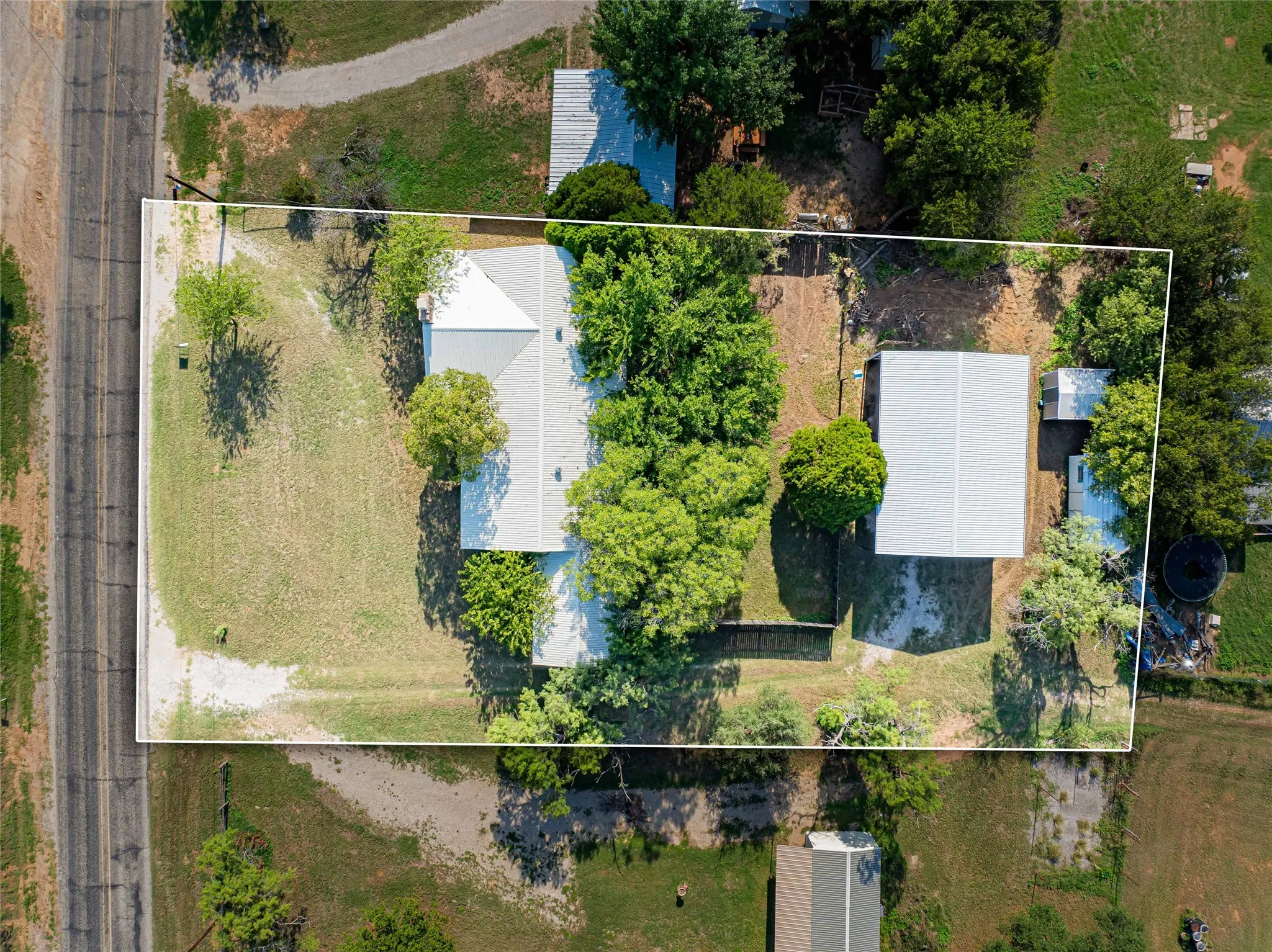 Aerial view of a property featuring a main residence with a light-colored roof, surrounded by mature trees