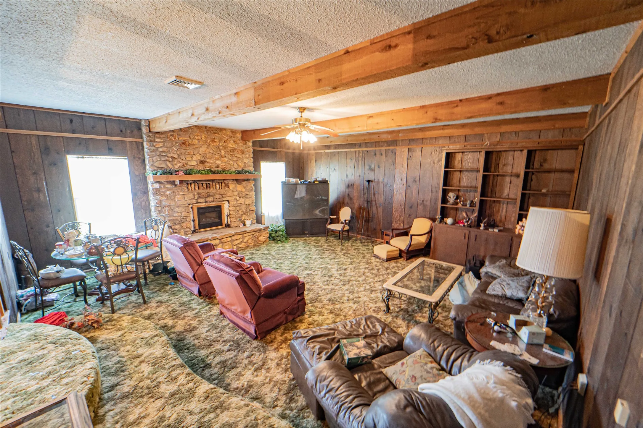 Spacious living area featuring a fireplace, exposed wood ceiling beams, wood paneling, built-in shelving, and multiple windows